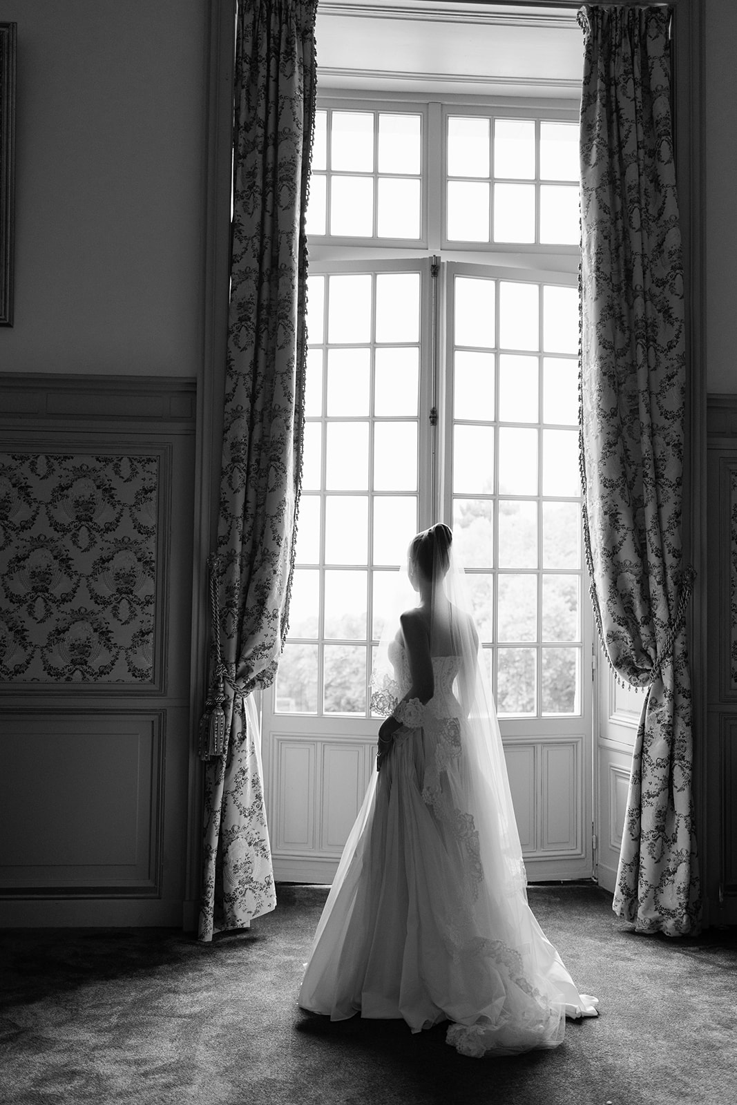 Black and white portrait of bride from behind looking through tall French window in chateau with lace gown and veil