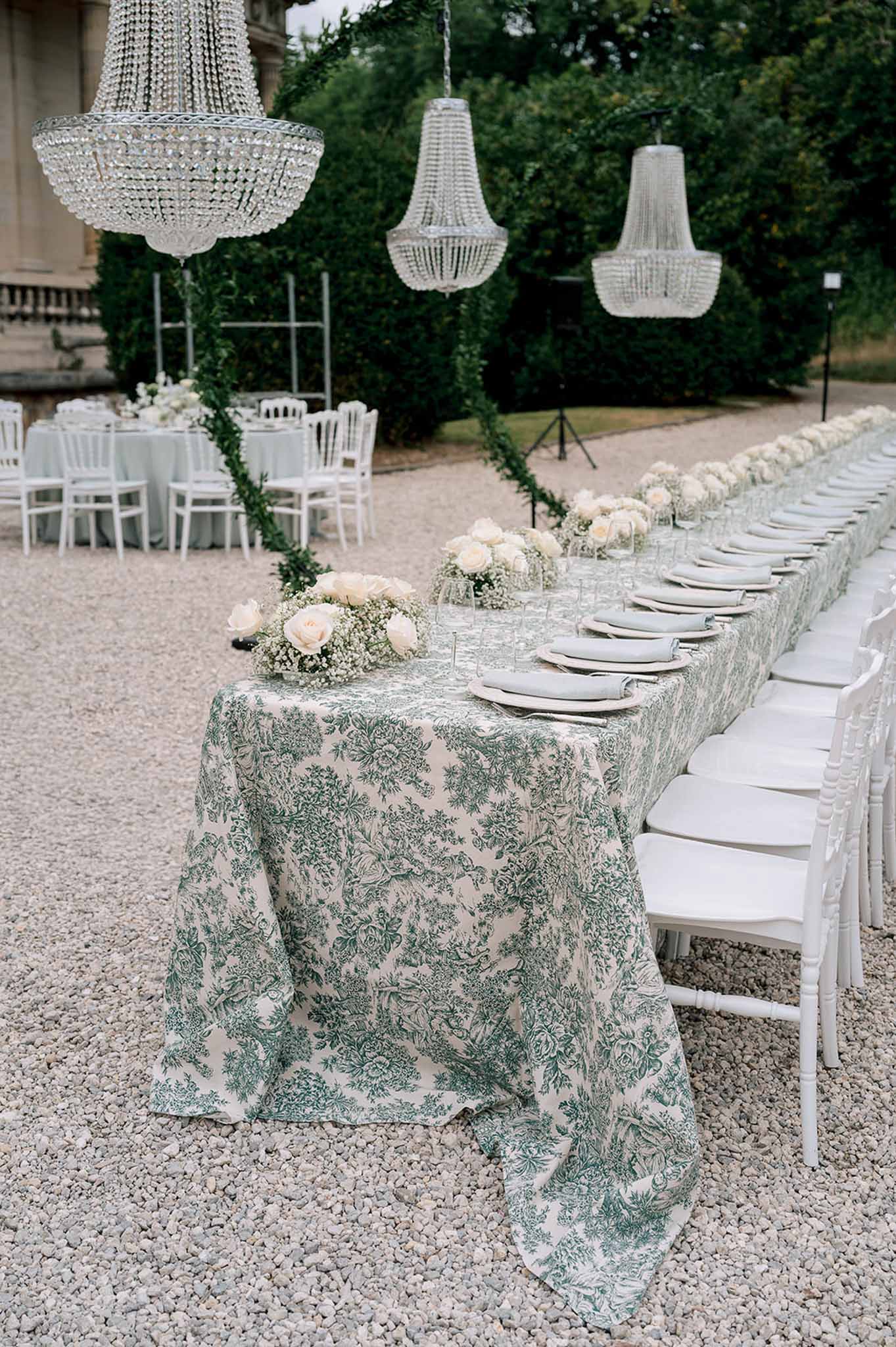 Long feasting table with toile de Jouy tablecloth, sage napkins, blush rose centerpieces, and crystal chandeliers on chate...