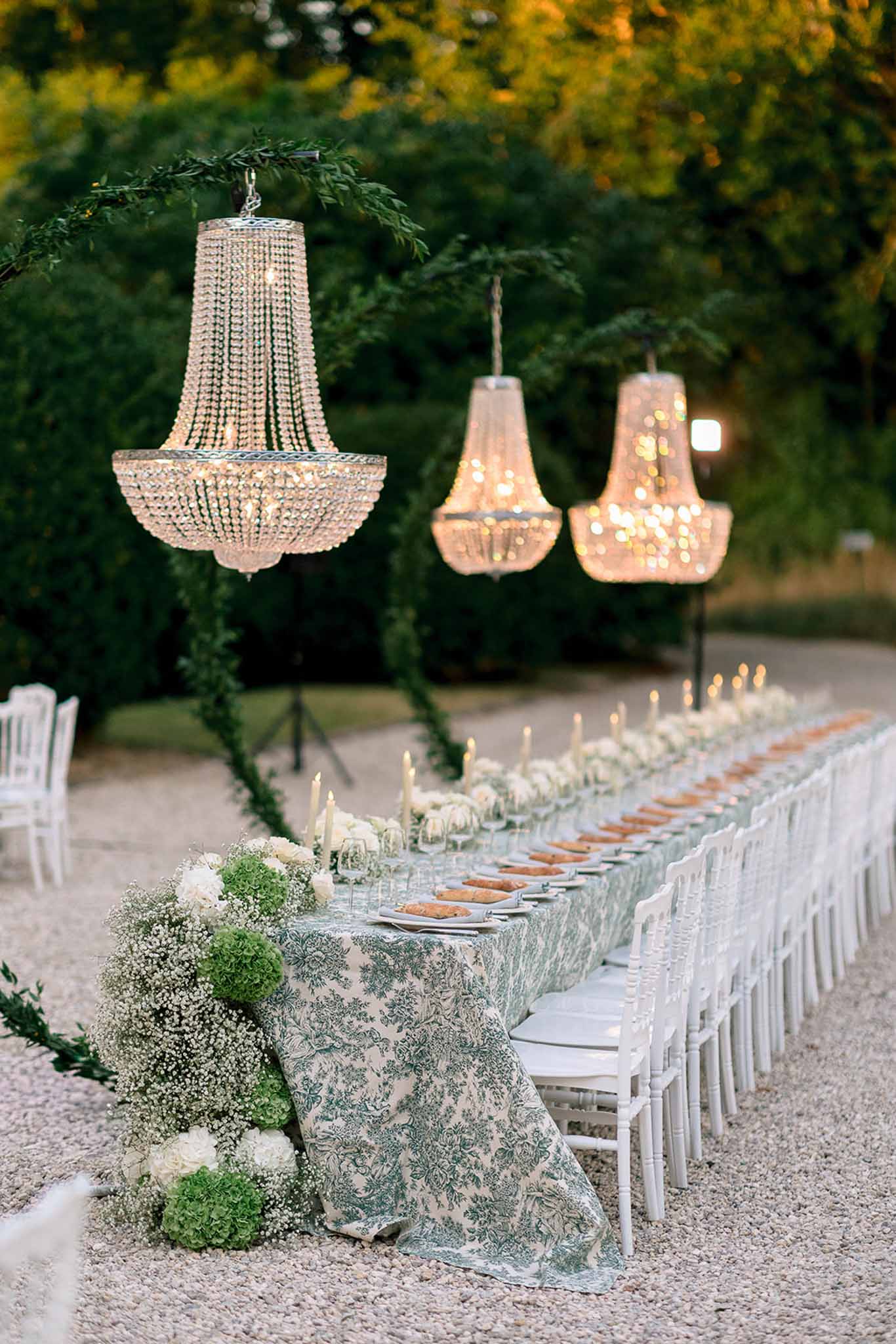 Long banquet table with green toile cloth, white roses, suspended chandeliers, and Chiavari chairs at dusk