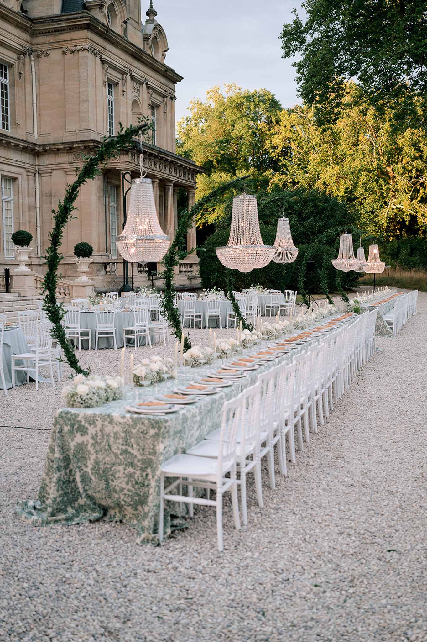 Outdoor reception at dusk with head table in sage green toile cloth, suspended chandeliers, and chateau facade