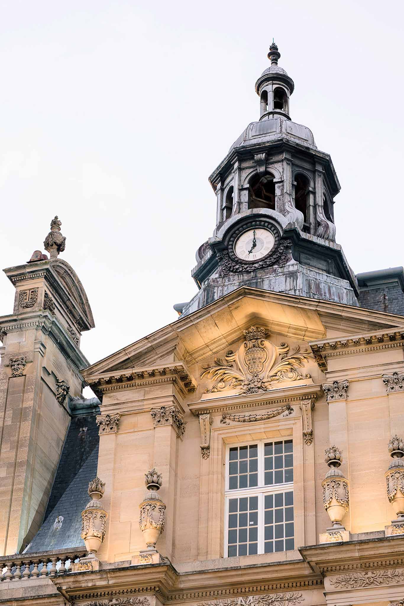 Uplit golden stone facade with carved urns, coat of arms cartouche, and oxidized clock tower belfry