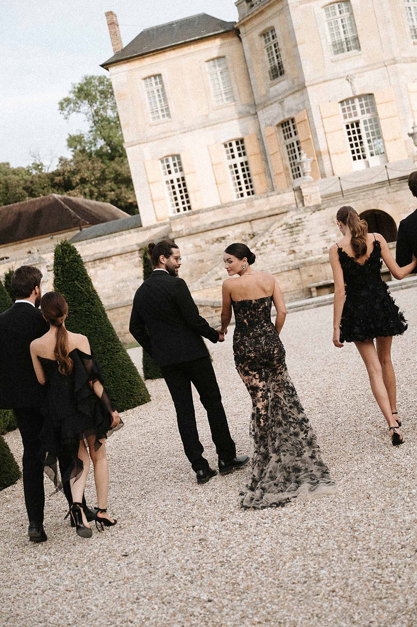 Wedding party in all-black attire walking toward French chateau, bride in black lace gown with train