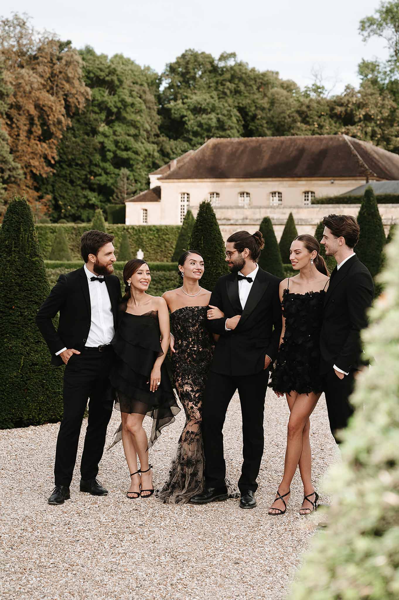 Six wedding guests in all-black formal attire pose on gravel path in French chateau gardens with topiary hedges