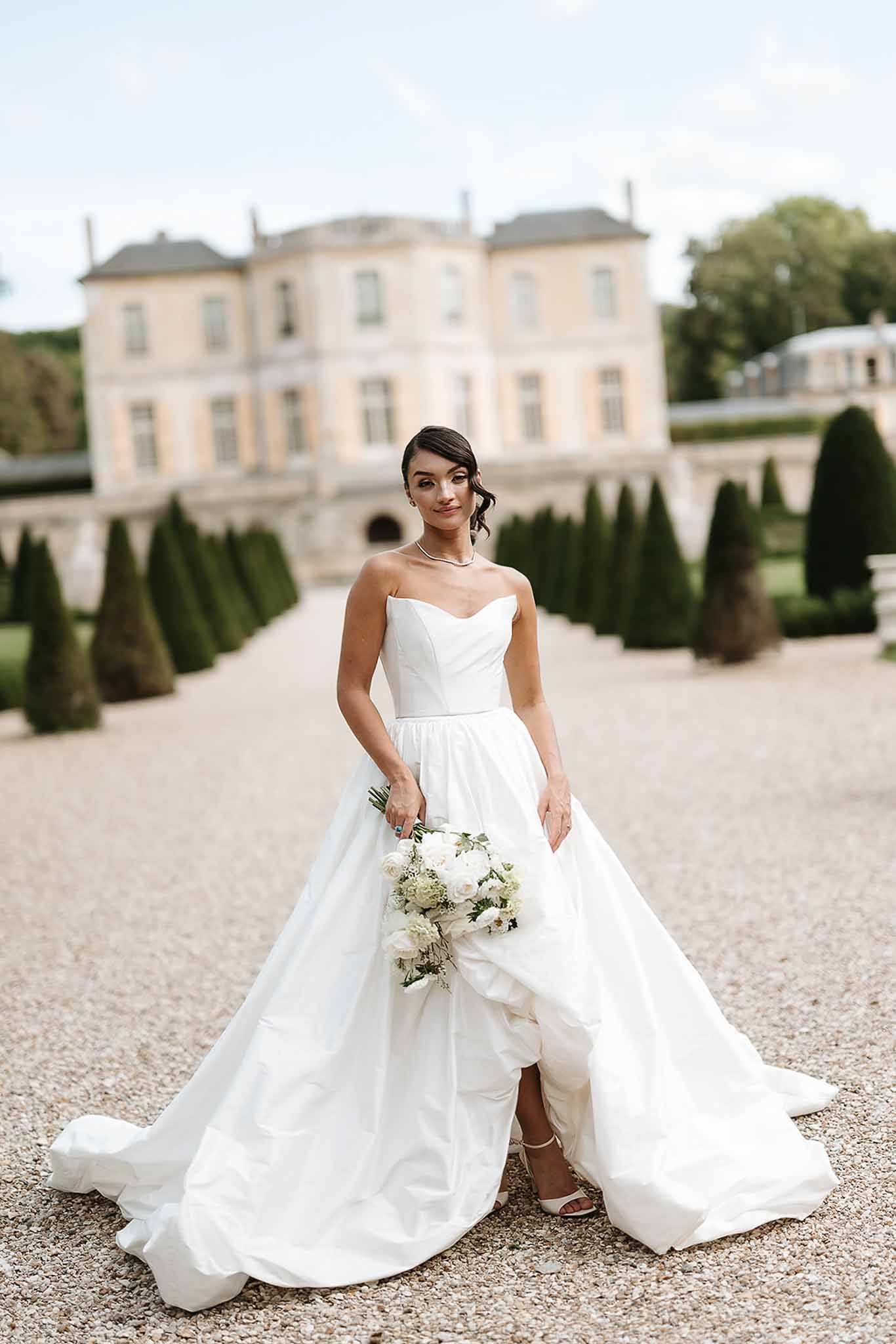 Bride in white strapless ball gown with full train holding white rose bouquet on chateau gravel driveway