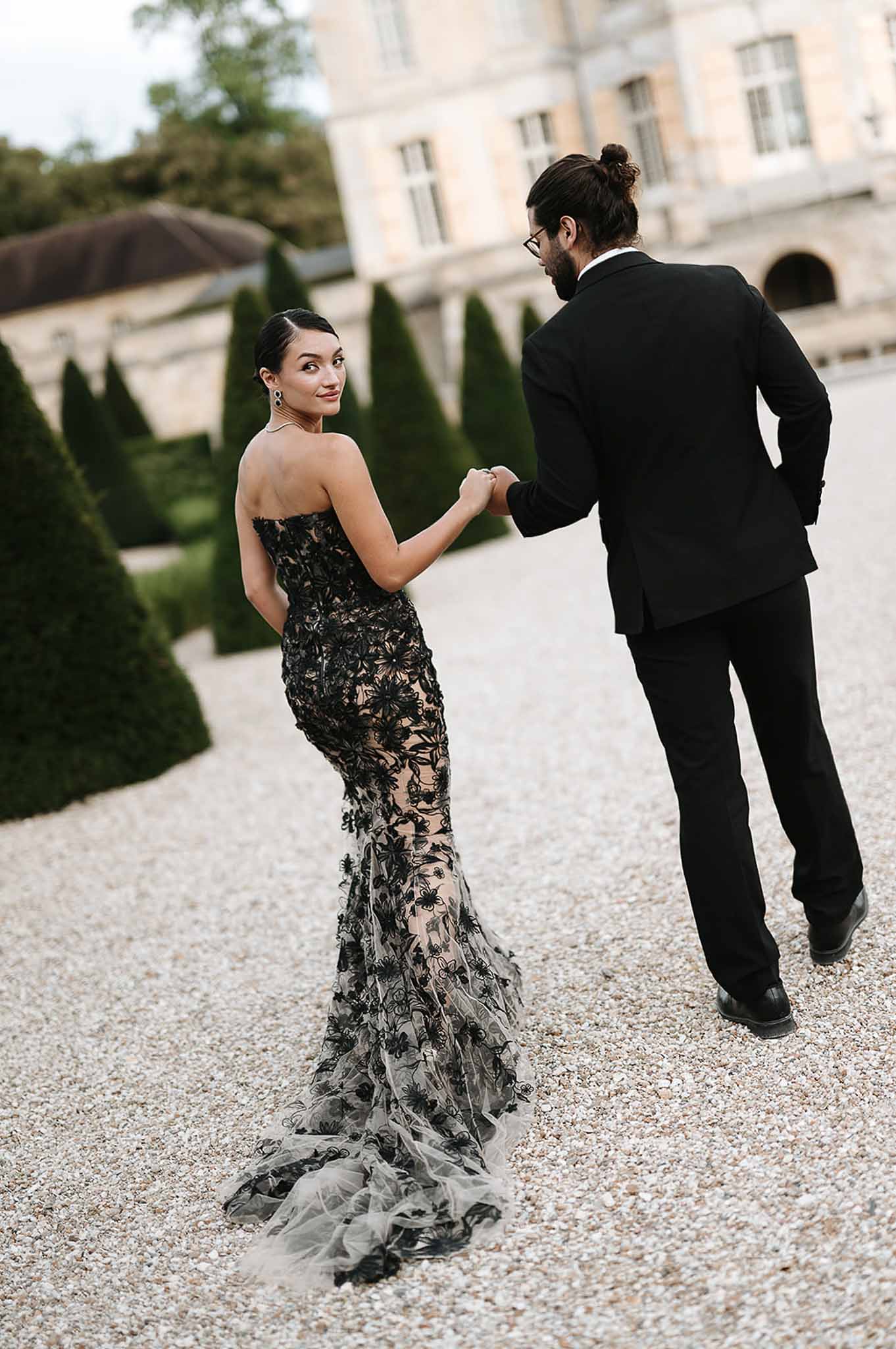 Bride in black floral lace mermaid gown and groom in black suit walking on chateau gravel forecourt