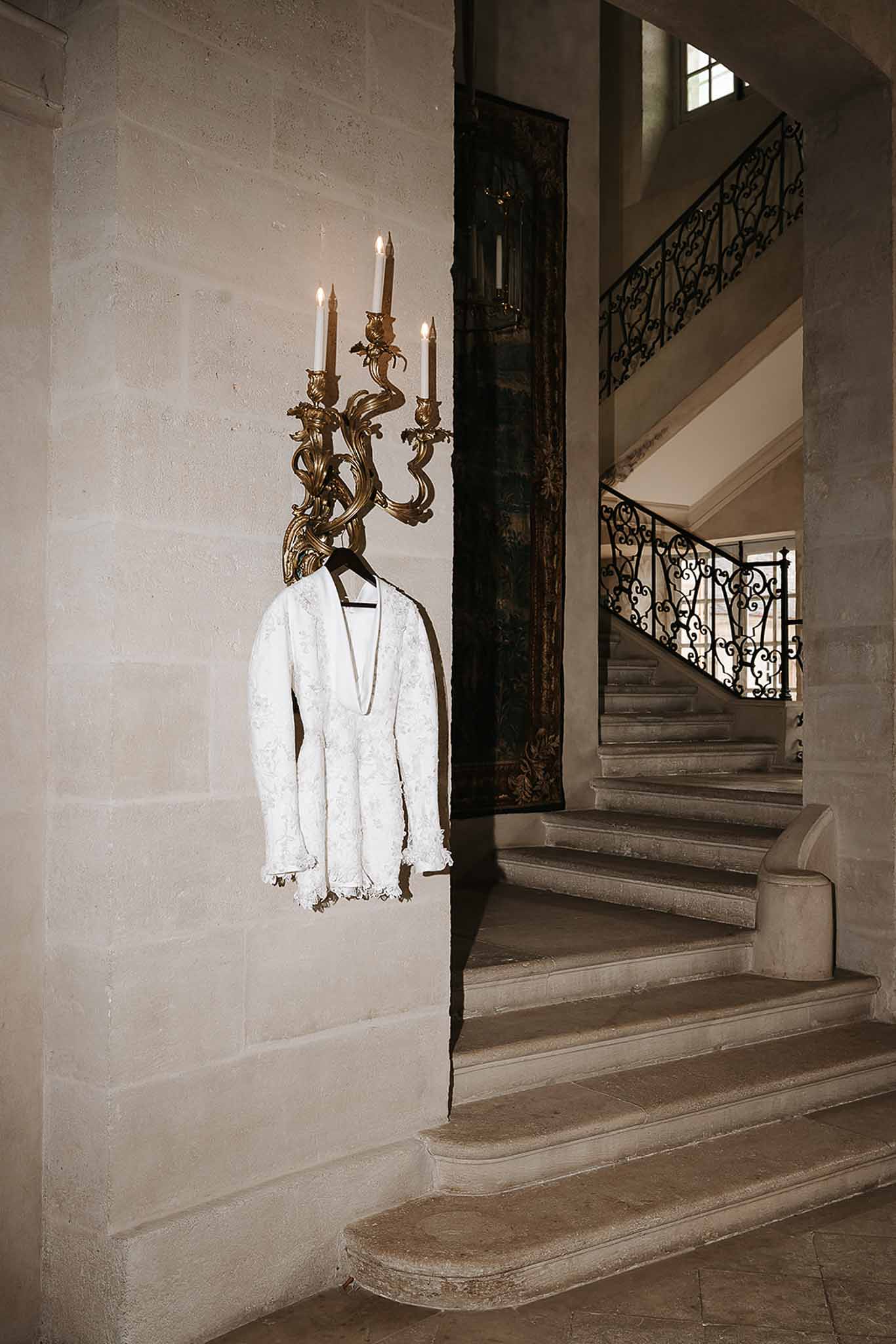 White lace bridal jacket hanging from a gilded baroque candelabra sconce in a chateau staircase hall