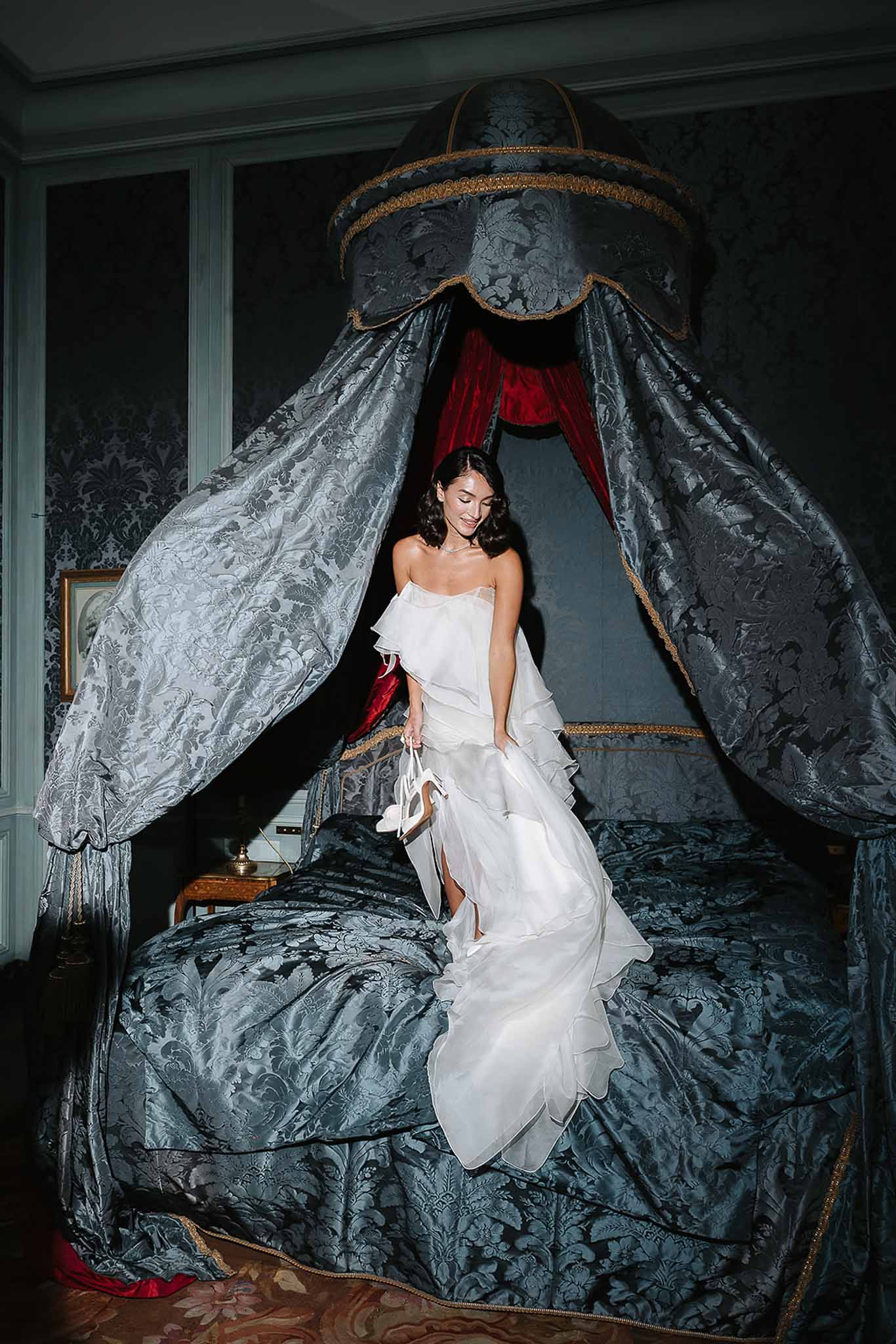 Bride in ruffled white gown standing on a canopied bed in an ornate chateau bedroom