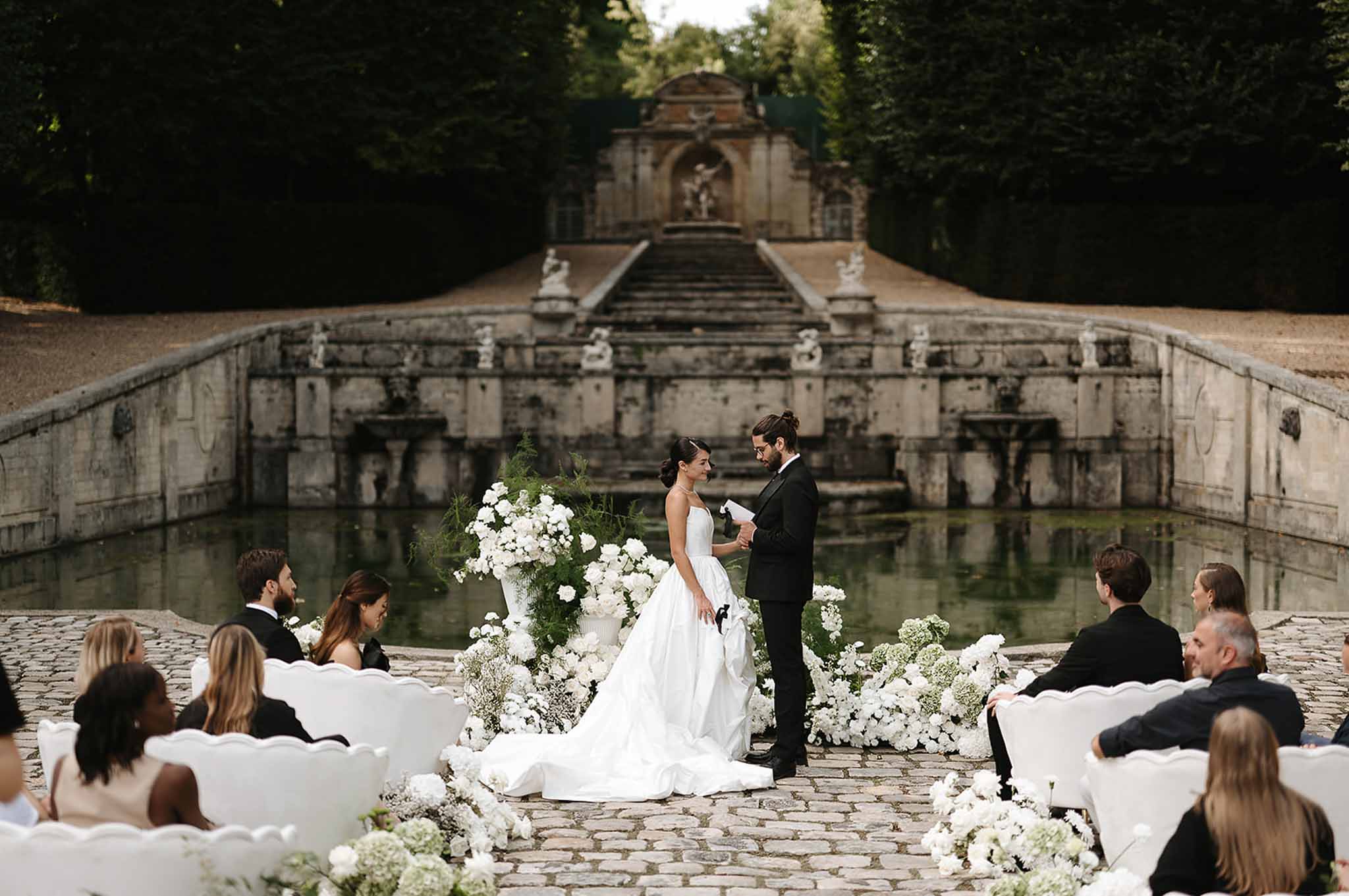 Ceremony before stone fountain with all-white peony urns, carved chairs, and formal garden staircase