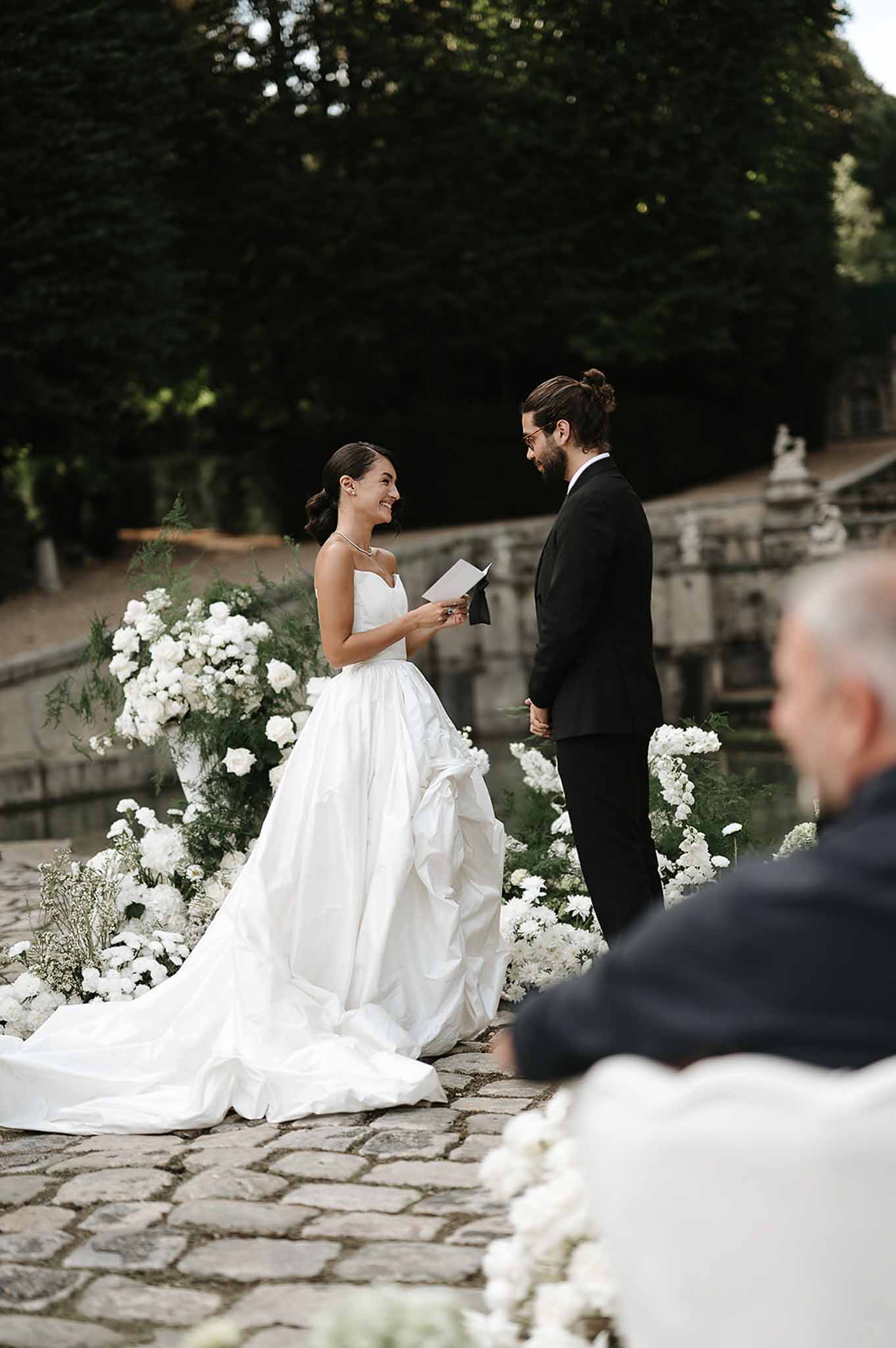 Bride reading vows to groom before white rose and fern ceremony backdrop on chateau terrace