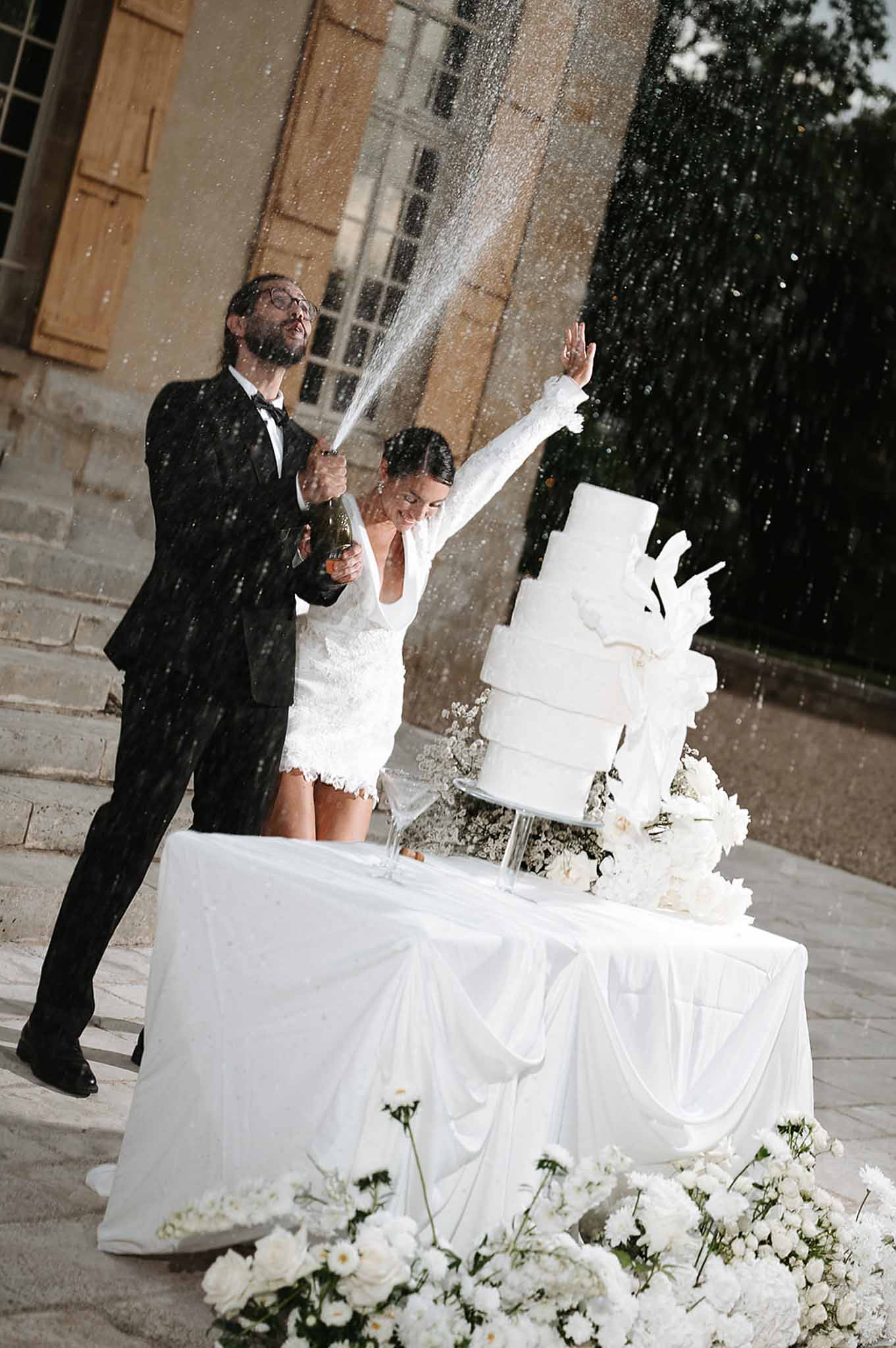 Bride and groom spraying champagne beside four-tier white wedding cake in front of stone chateau steps