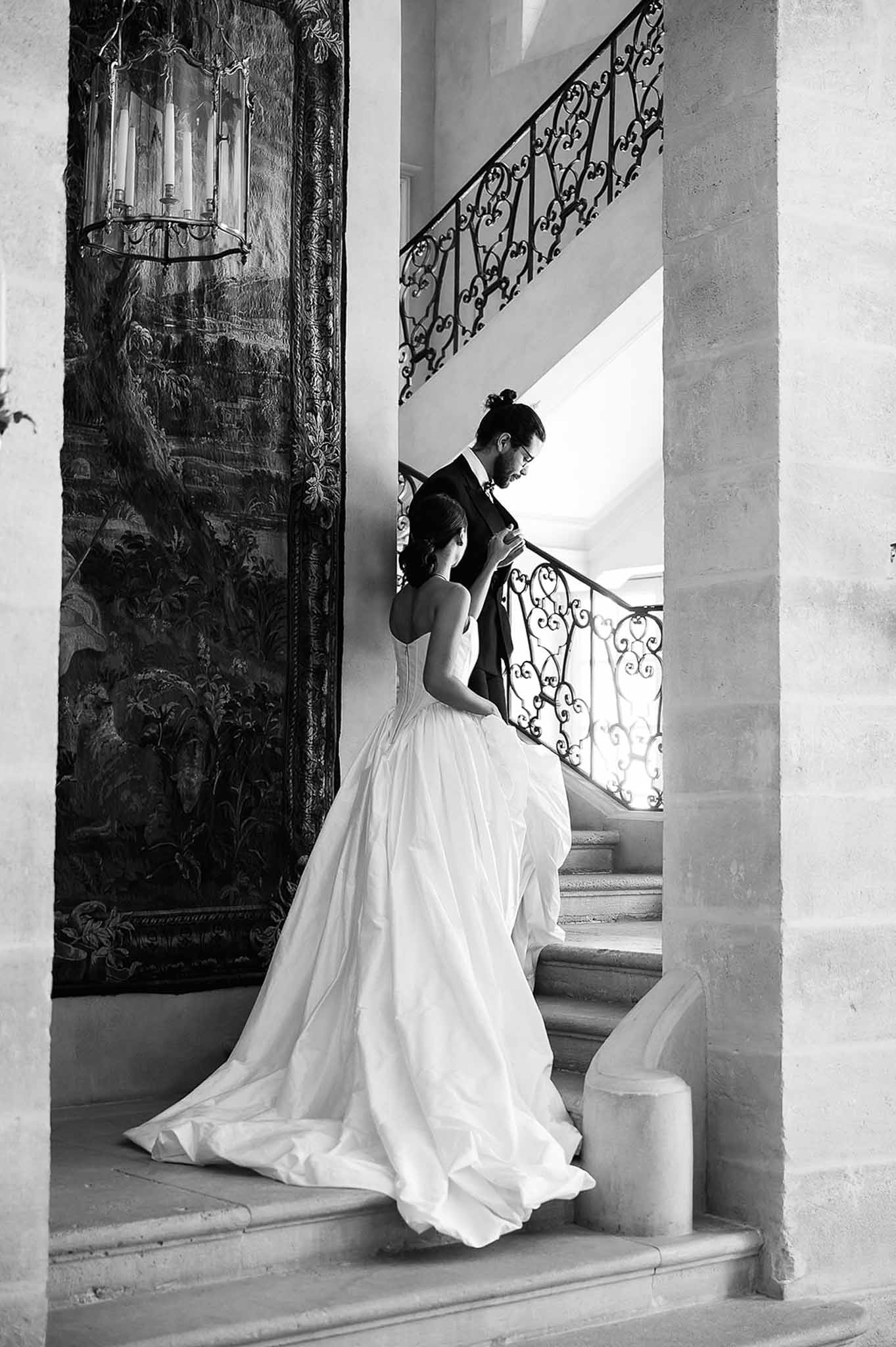 Black-and-white portrait of groom fastening bride's ballgown on grand stone staircase with iron balustrade