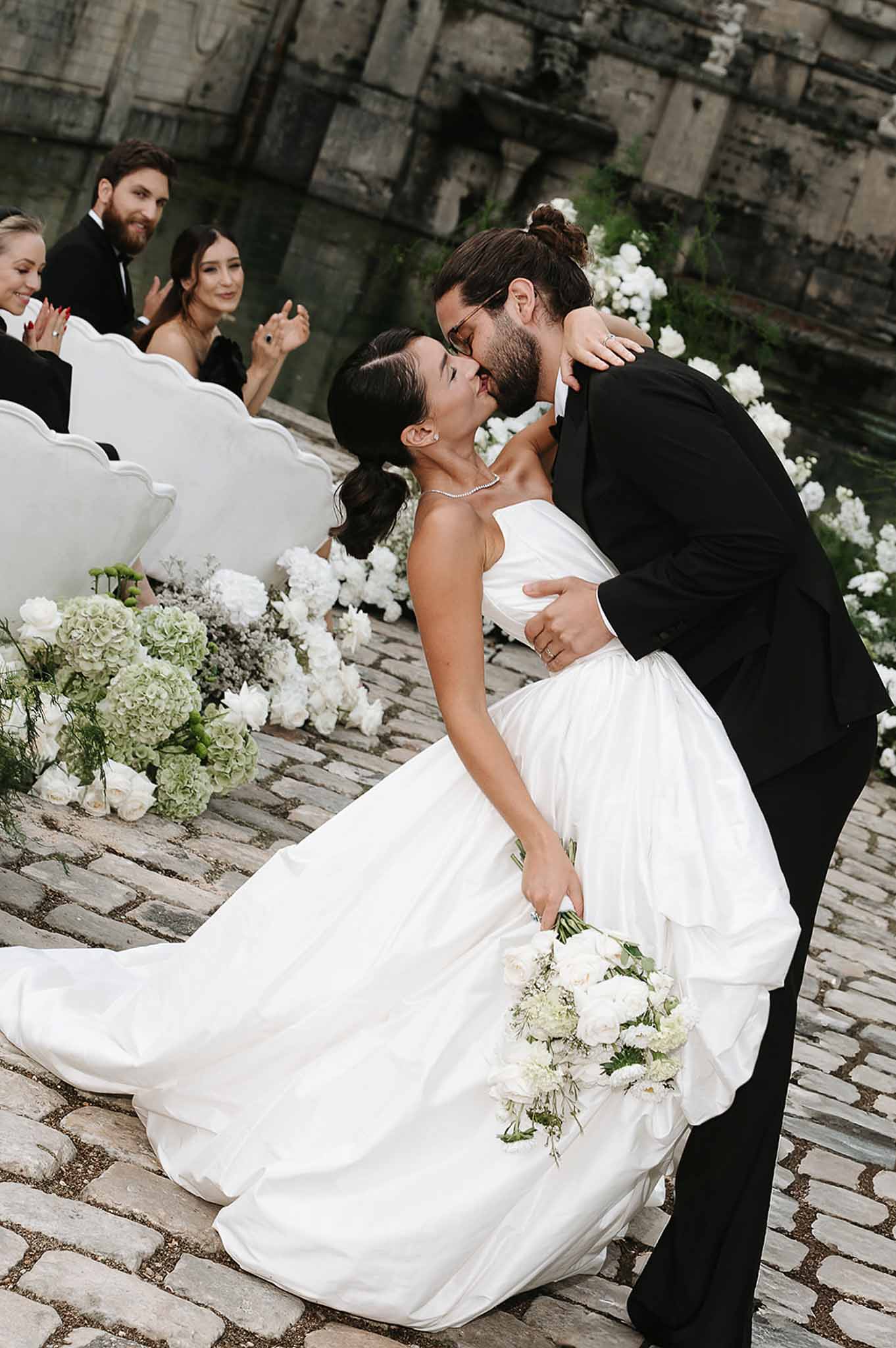 First kiss on cobblestone with white hydrangea and rose aisle arrangements against aged stone wall