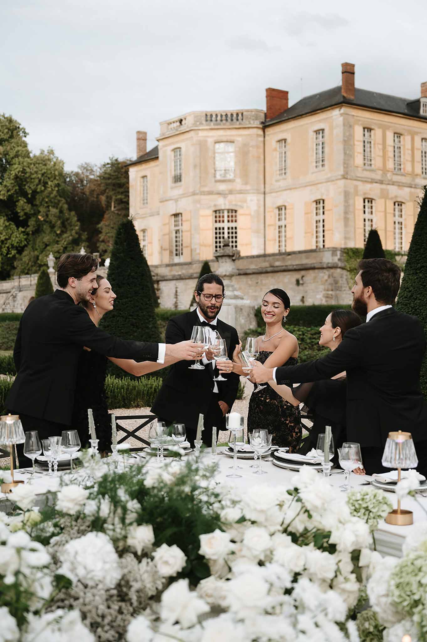 Six guests in black toasting at white rose and hydrangea table with brass lamps before chateau garden