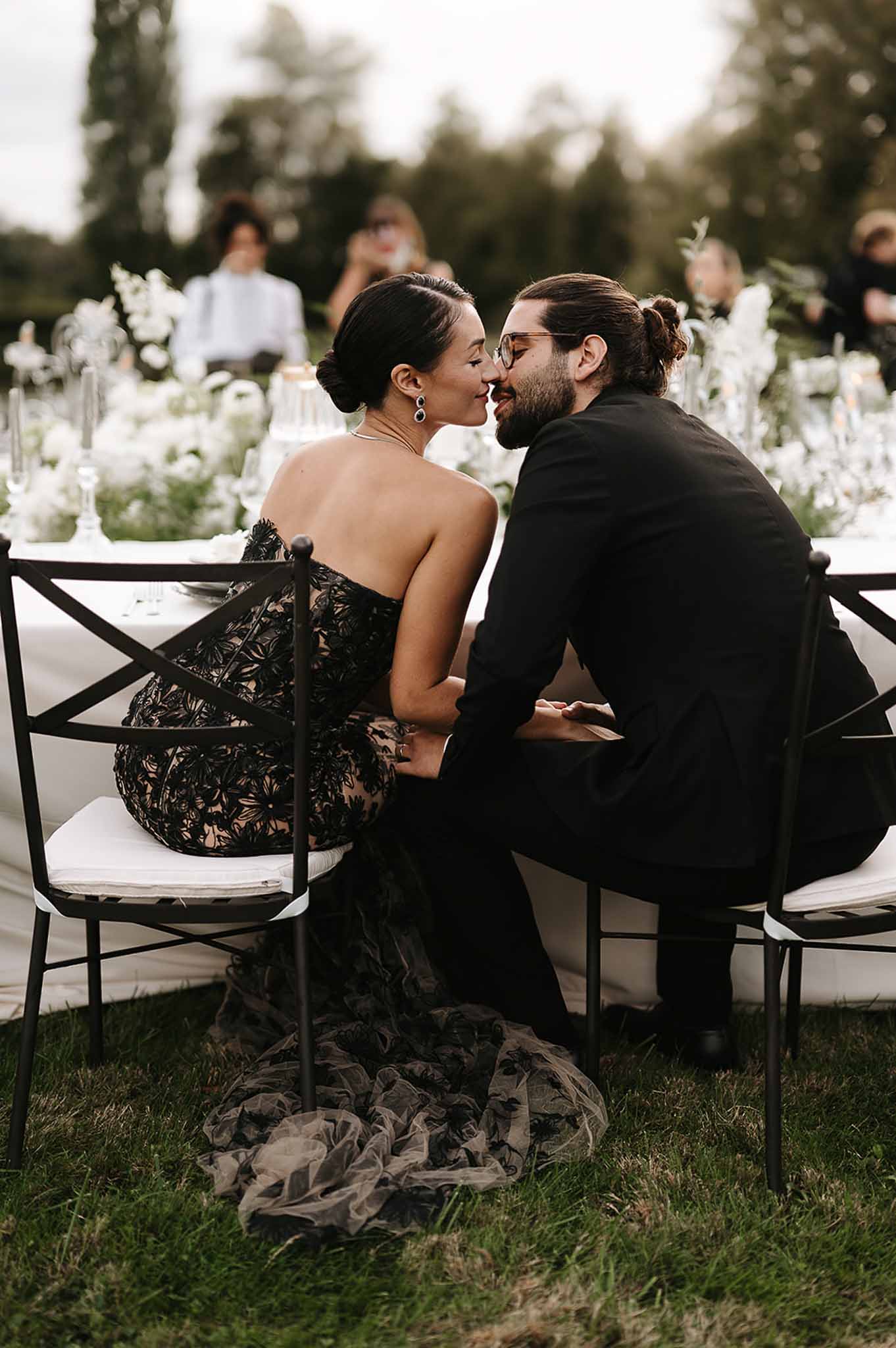 Couple kisses at sweetheart table with bride in black lace gown and trailing tulle train on grass