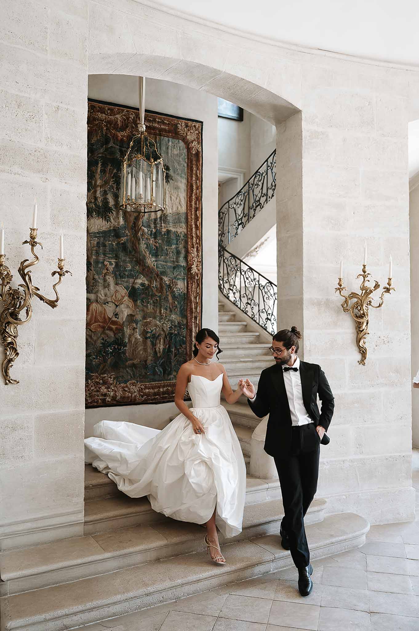 Bride in ivory ball gown and groom in black tuxedo descending grand staircase beneath gilded tapestry and lantern