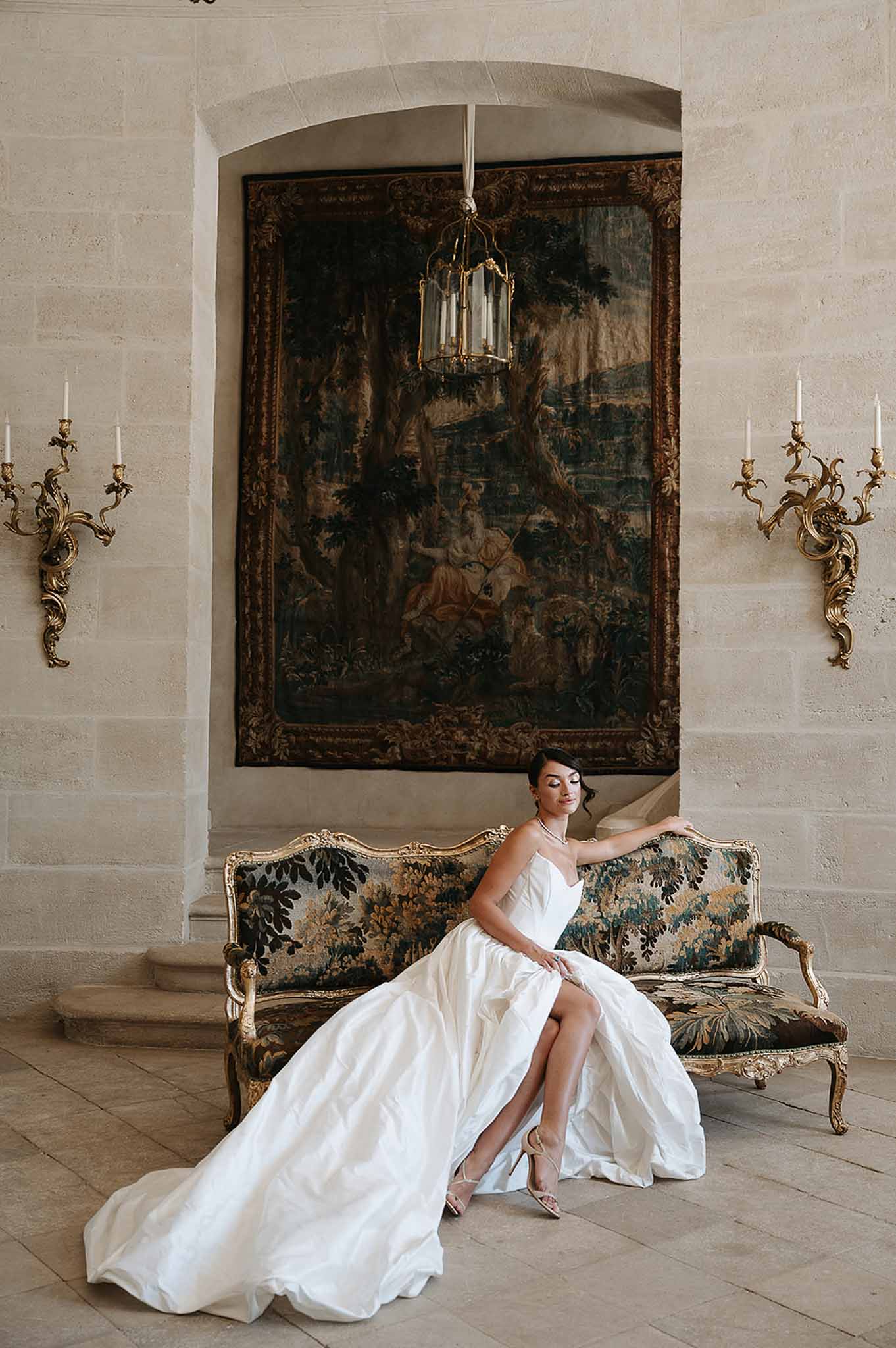 Bride in strapless ball gown seated on green tapestry canape beneath verdure tapestry and sconces