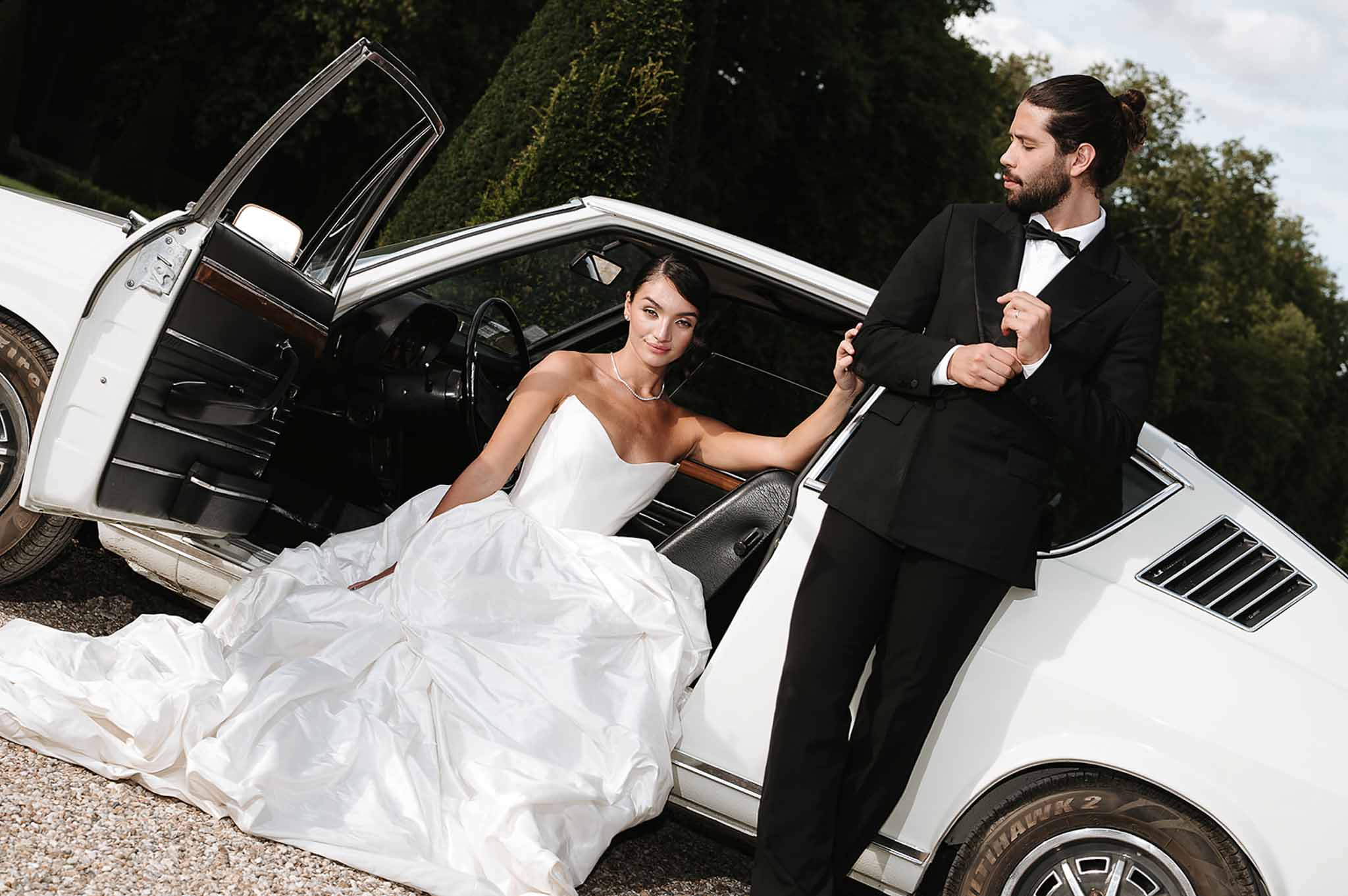 Bride in strapless ballgown seated in gull-wing vintage car as groom in tuxedo stands beside