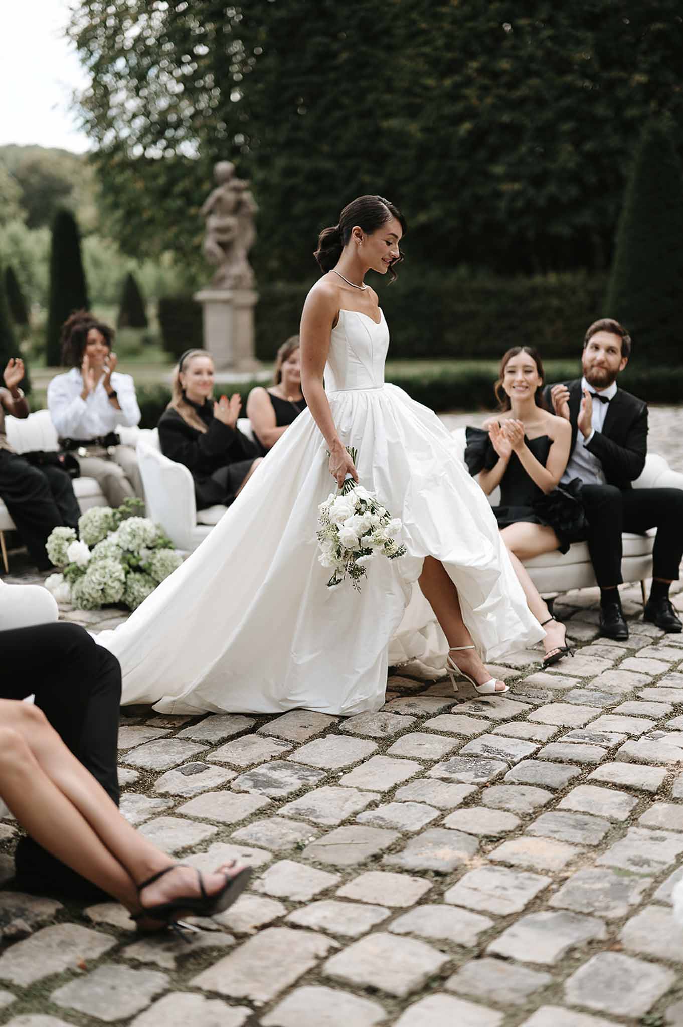 Bride in white ballgown walks cobblestone aisle past all-black dressed guests with white hydrangea clusters