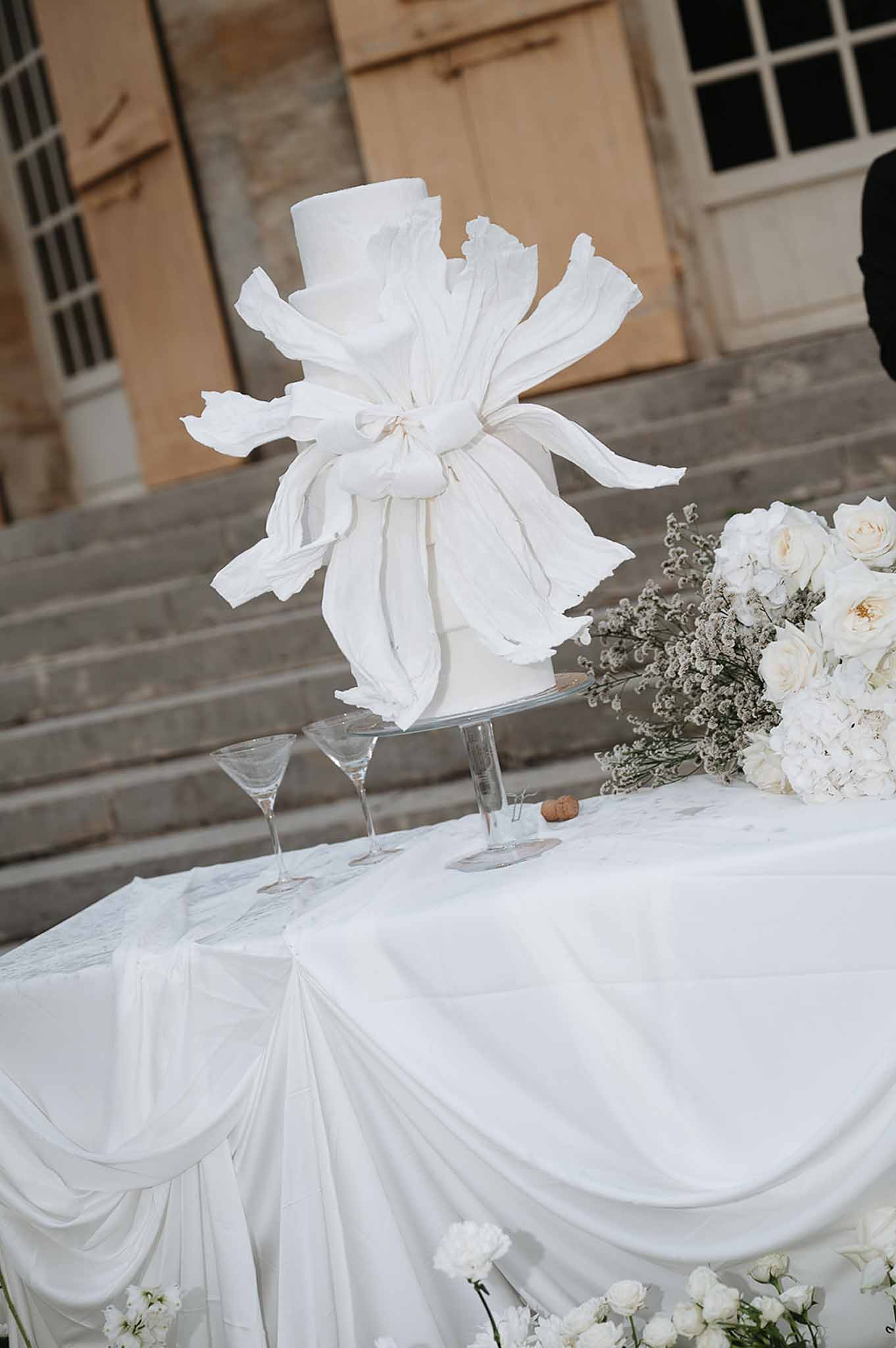 Two-tier white cake with sculptural sugar ruffles on draped table with white roses and hydrangeas