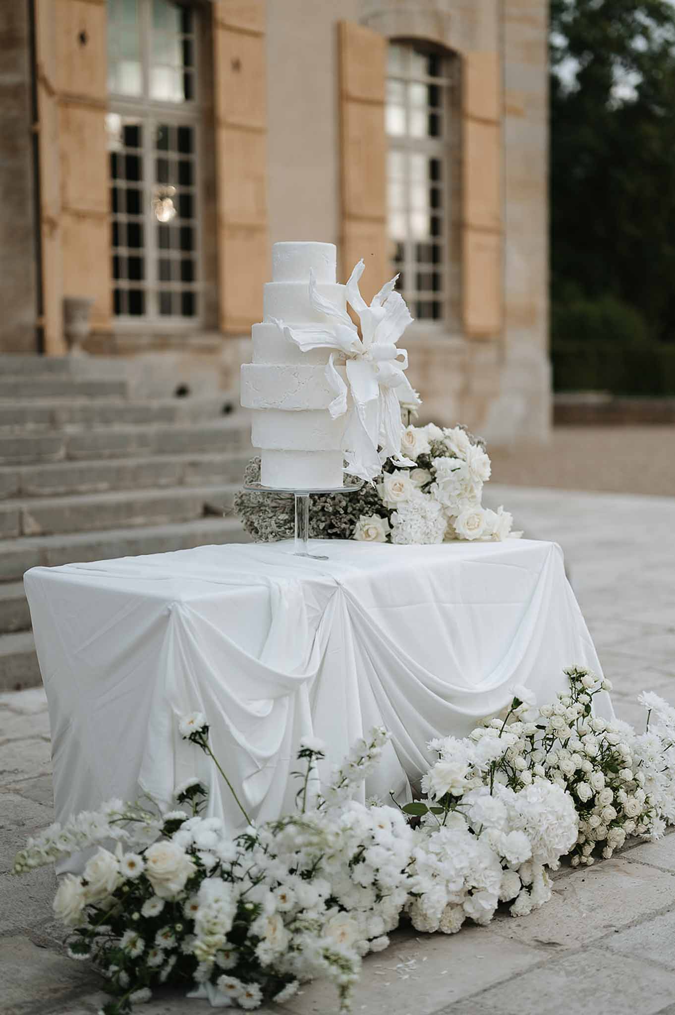 Five-tier white ribbon-bow cake on draped table with white rose ground arrangement before chateau steps