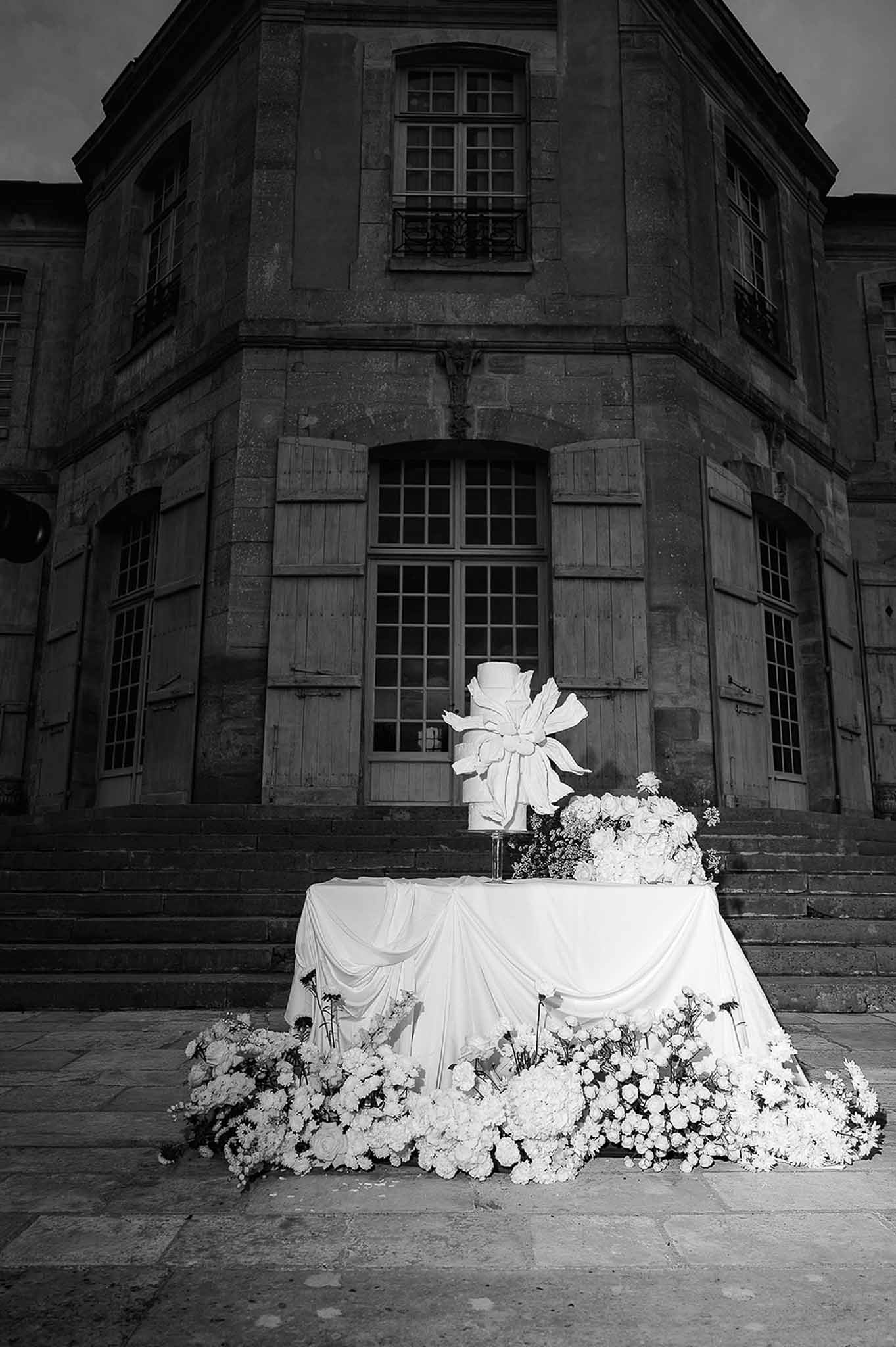 Black and white wedding cake on draped table with ground-level rose arrangements before chateau steps