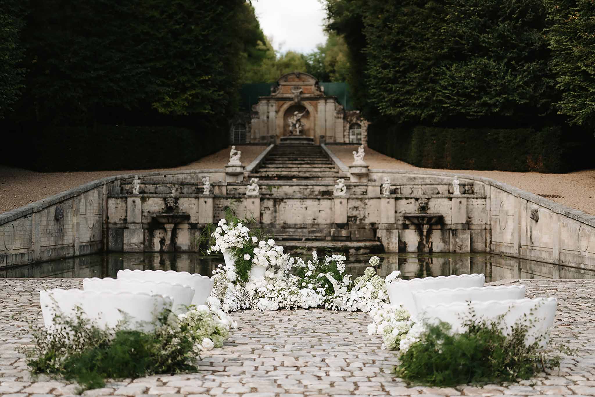 All-white floral altar installation on cobblestones before grand stone cascade staircase with statues
