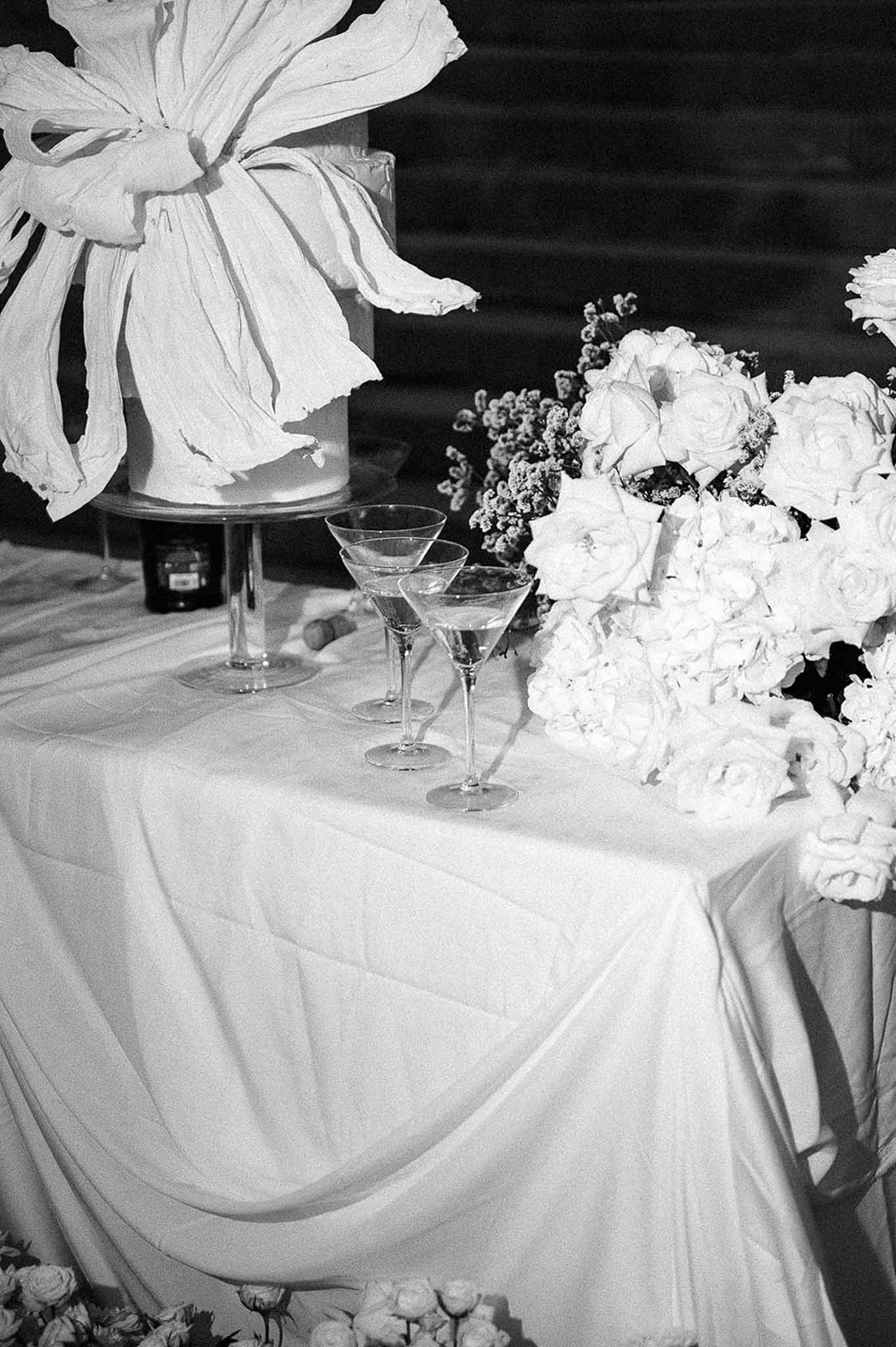 Black and white of tiered cake with fabric bow, champagne coupes, and dense rose arrangement on draped table