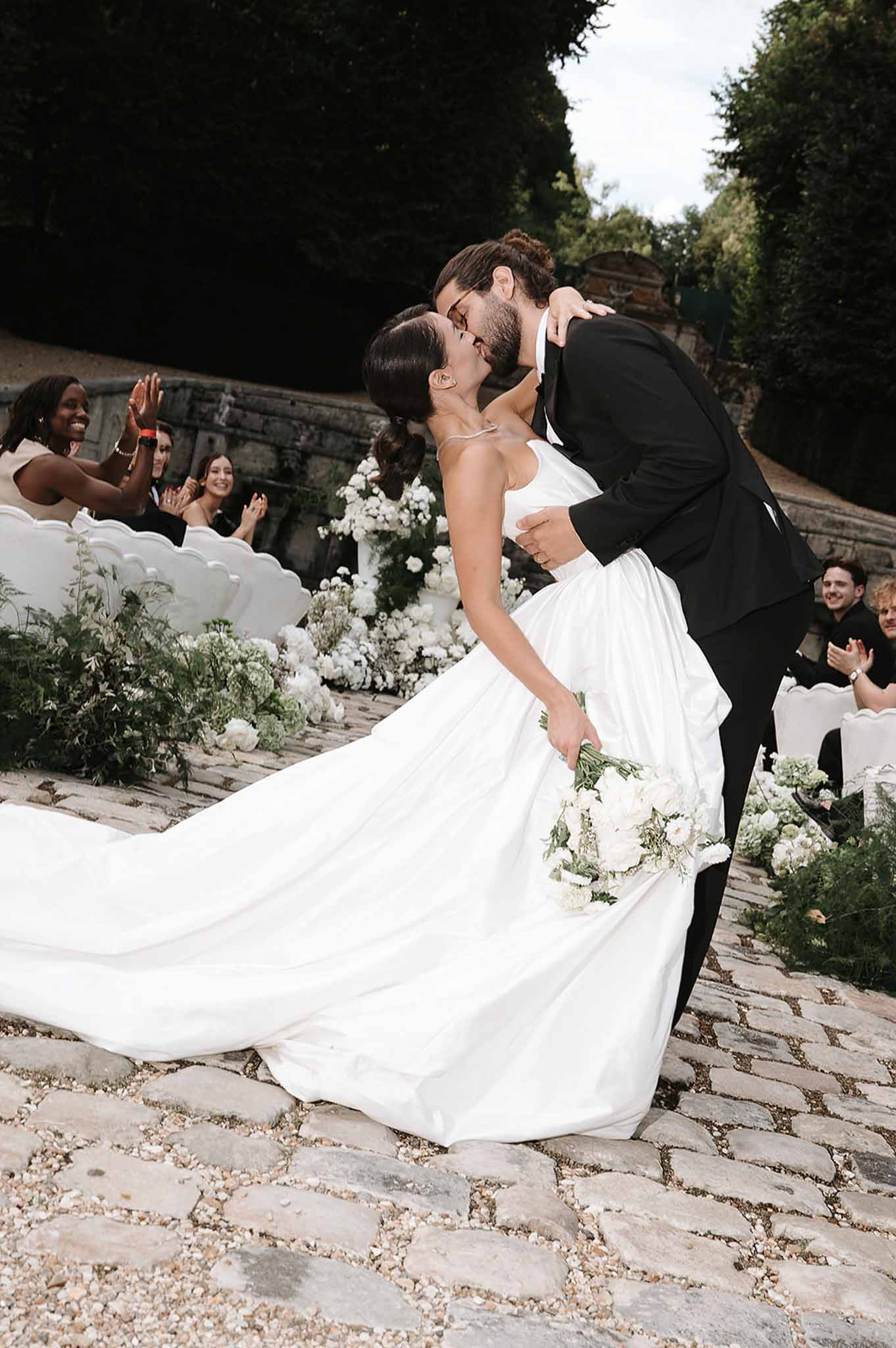 First kiss with groom dipping bride on cobblestone aisle lined with white roses and greenery