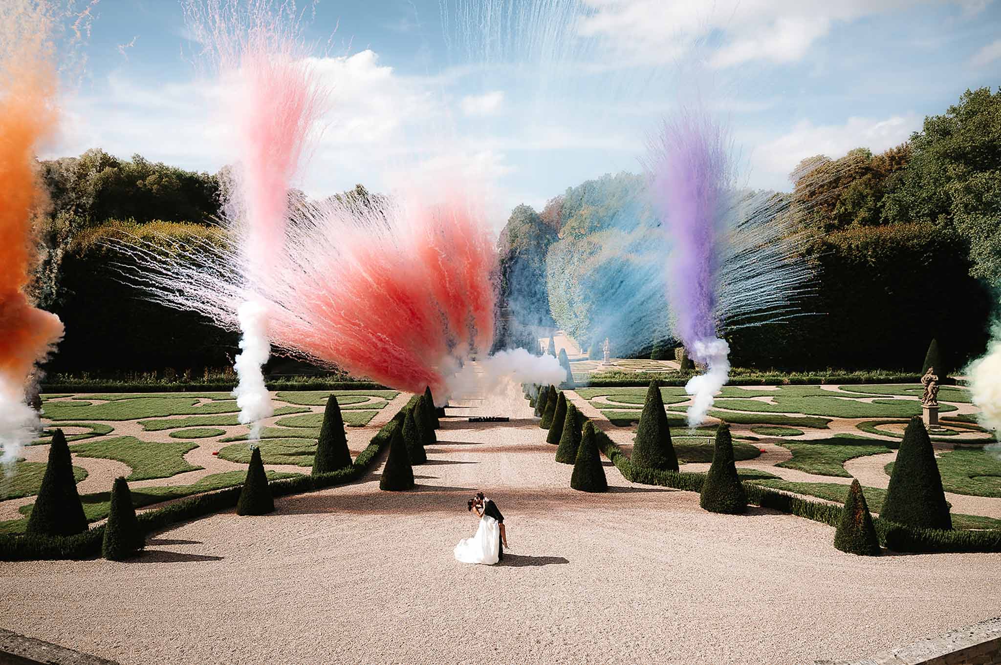 Couple dip kiss in formal French garden with coloured smoke cannons arcing over topiary allee and parterre hedging