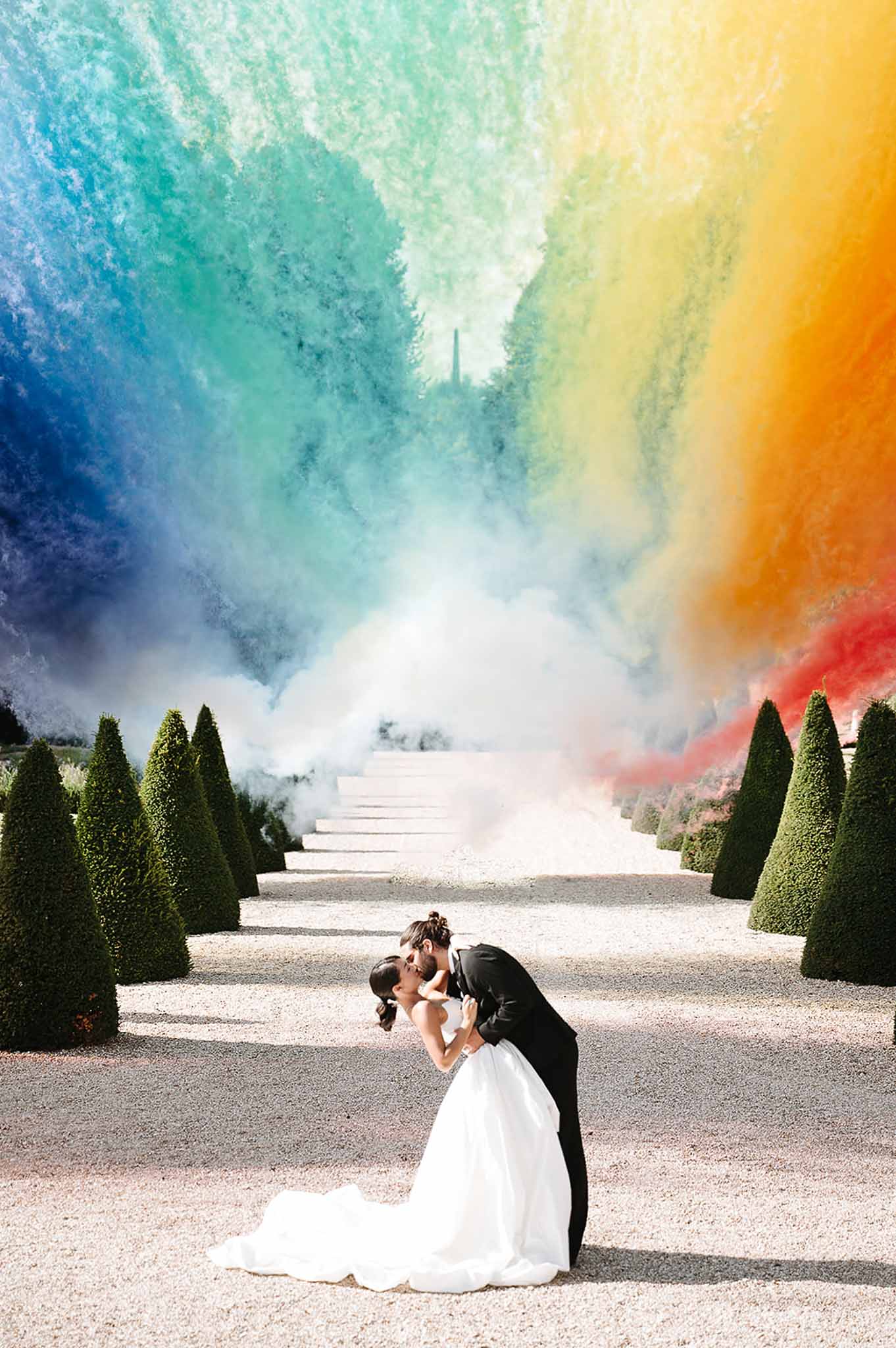 Groom dips bride for a kiss in formal garden as rainbow smoke bombs billow above topiary-lined allee