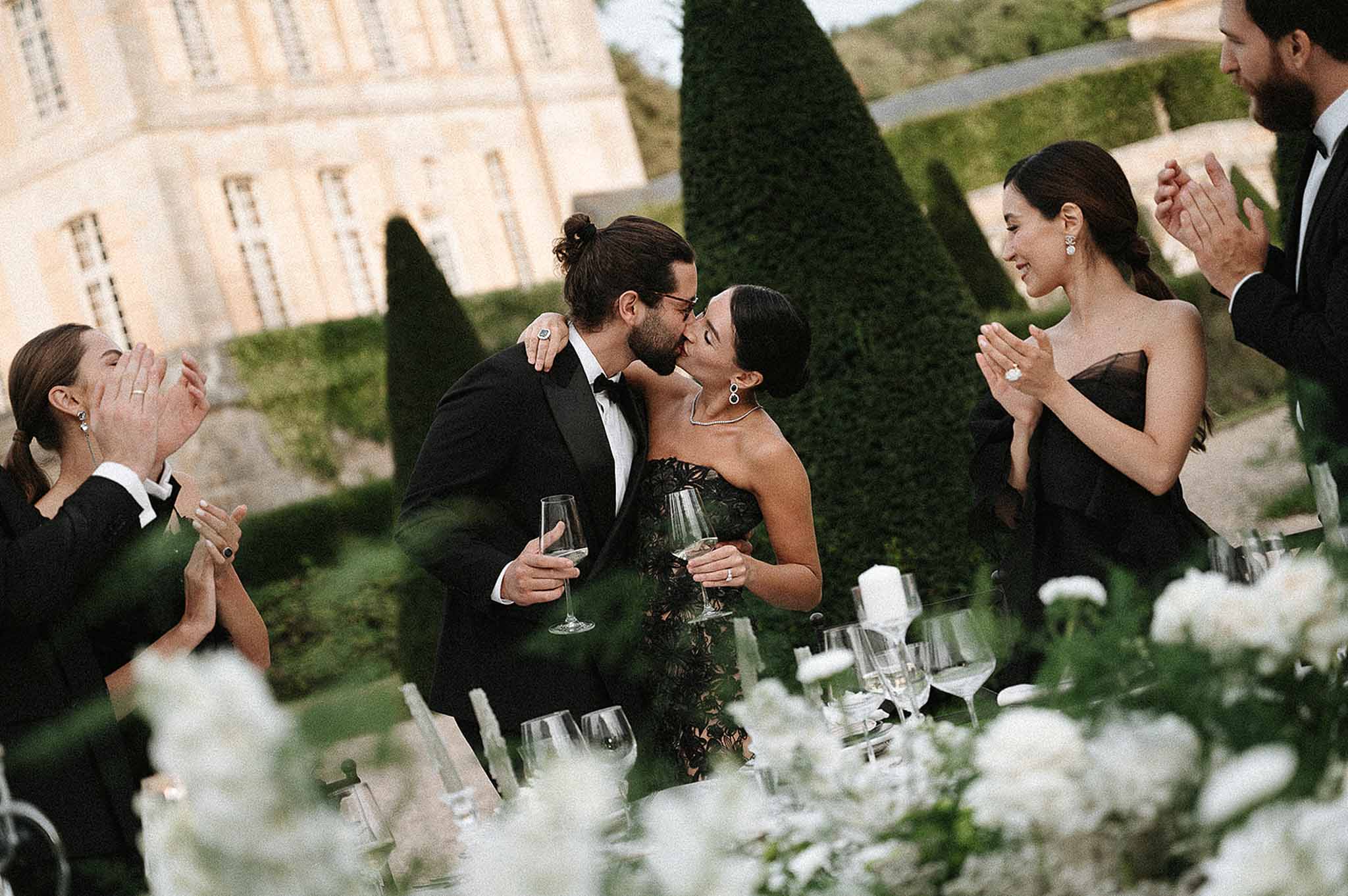 Couple kissing with champagne at all-black dress code reception with white rose table before chateau topiary