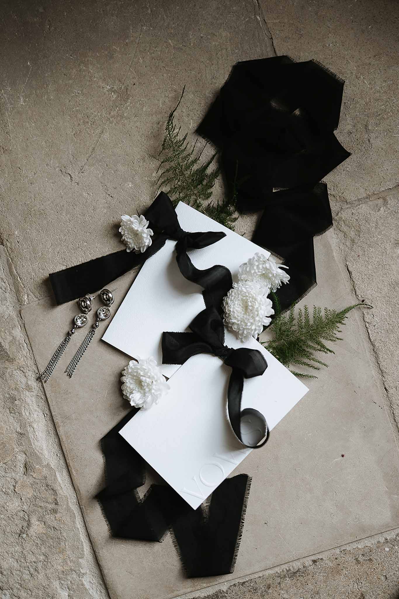 Monochrome flat lay with white cards tied in black ribbon, chrysanthemums, fern, and crystal drop earrings
