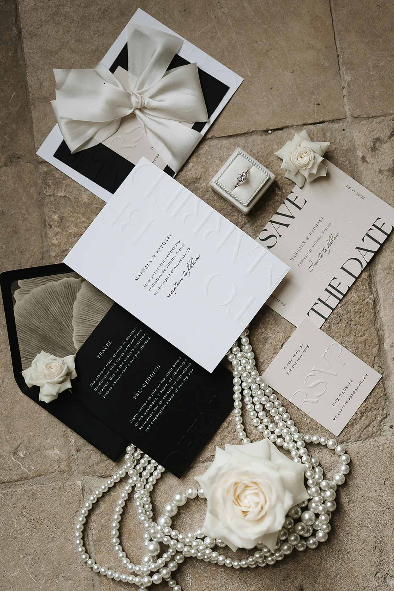 Wedding stationery flat lay with black and white invitations, pearl necklaces, cream roses, and velvet ring box
