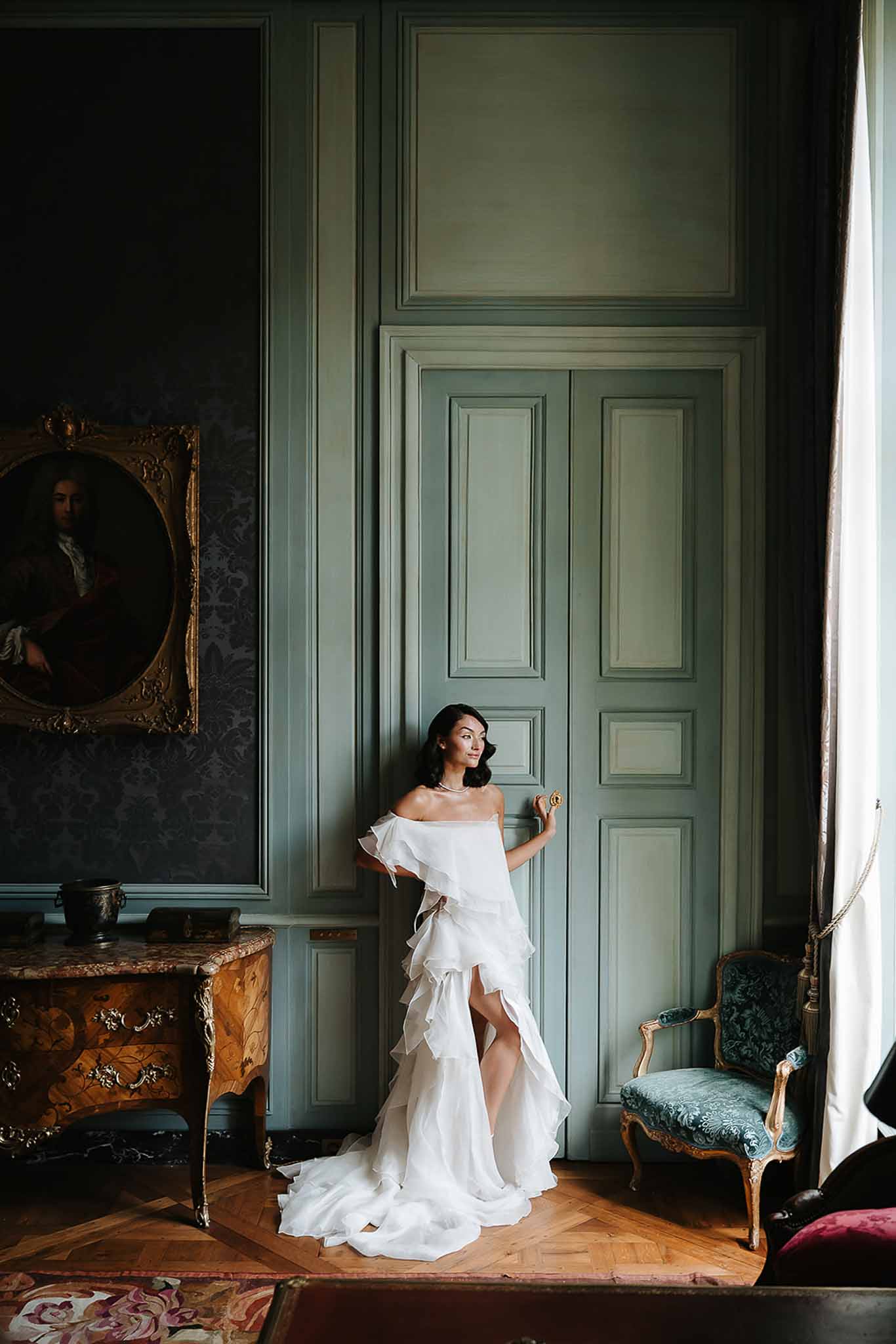 Bride in off-the-shoulder ruffled white gown leaning against sage-green paneled doors in a chateau room