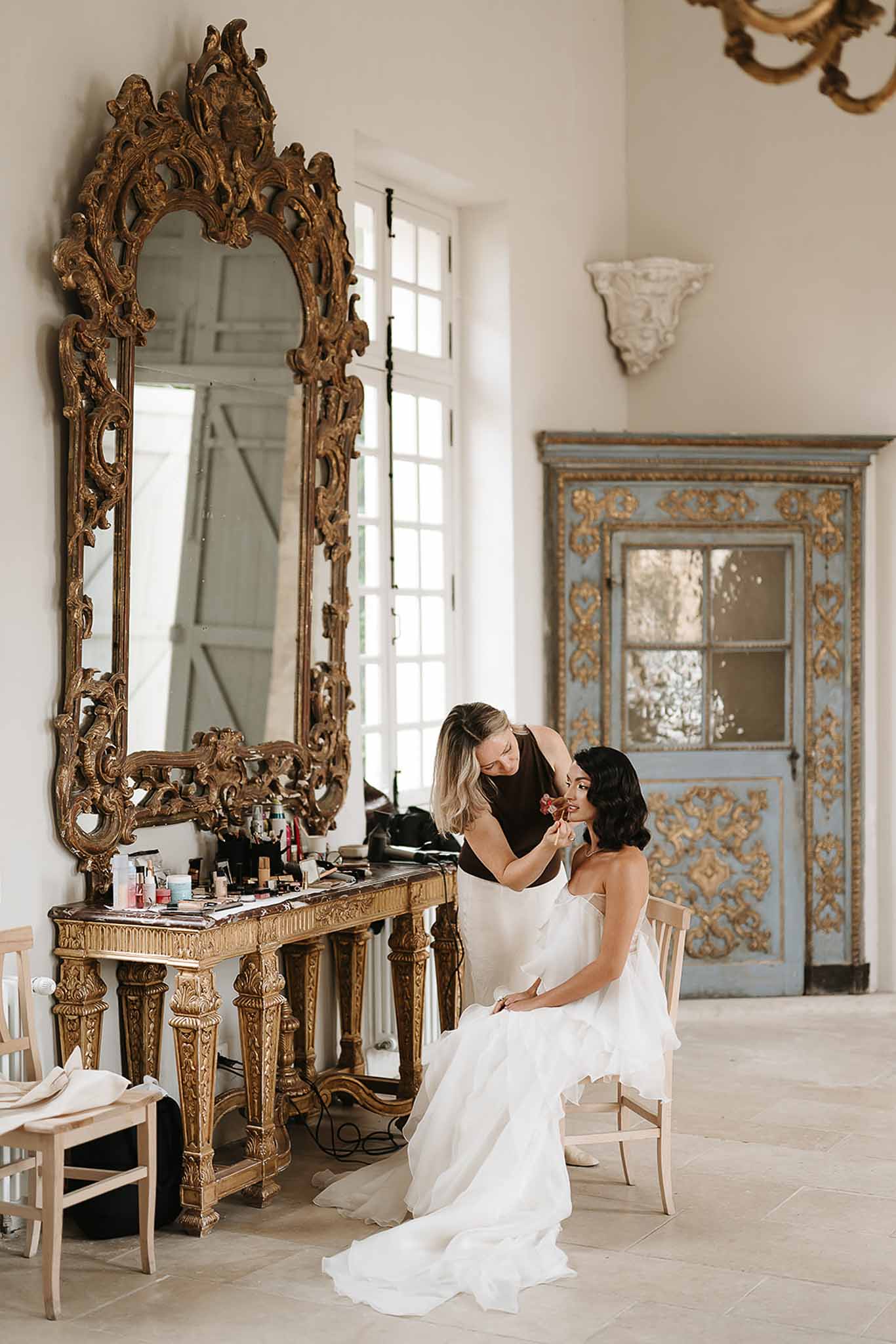 Bride in strapless ruffled gown having makeup applied beside gilt mirror and blue antique armoire in chateau room