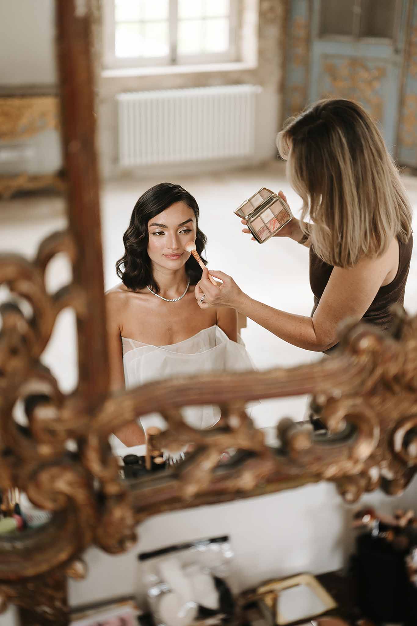 Bride having makeup applied reflected in ornate gold-framed baroque mirror inside chateau room