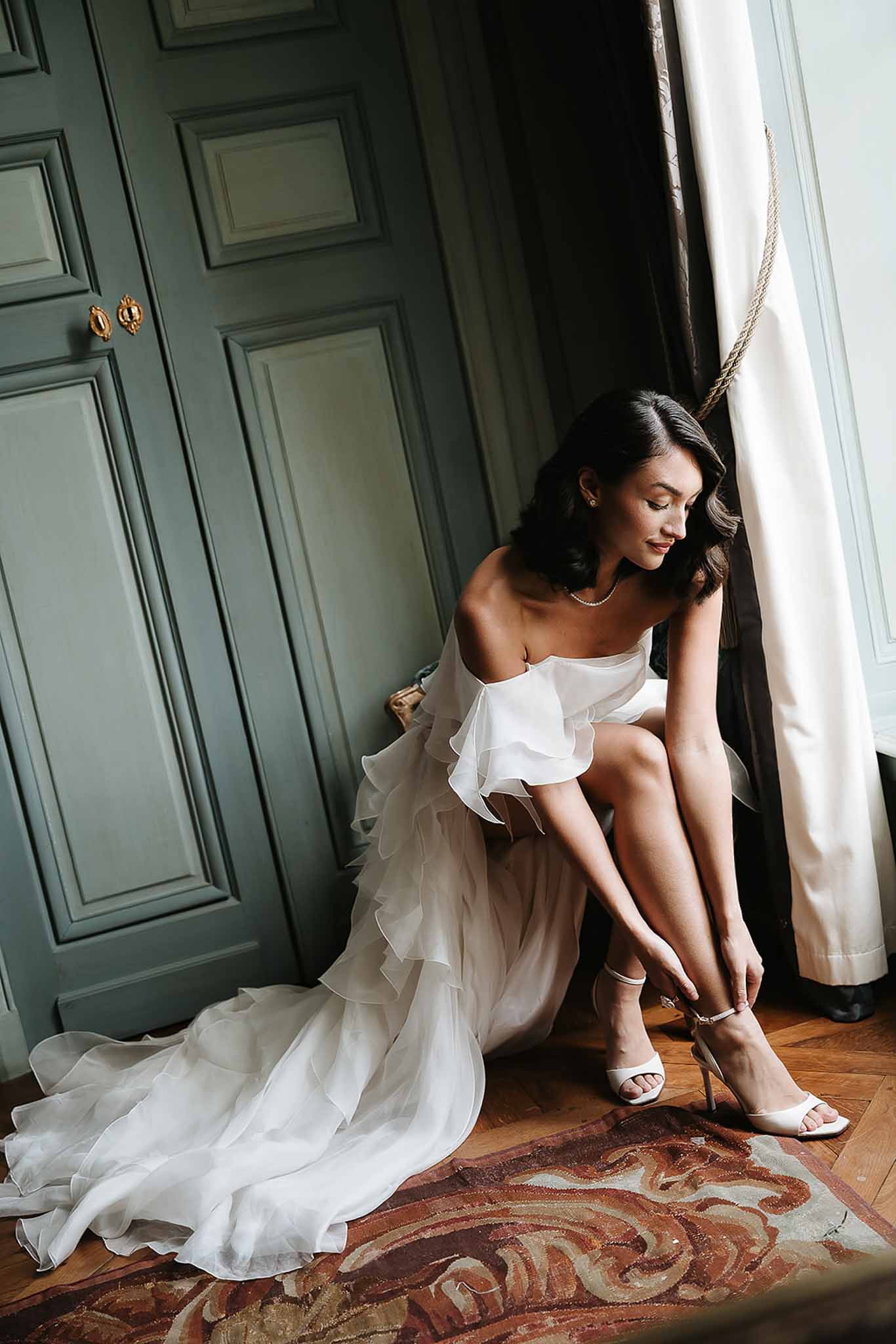 Bride fastening white satin heel by tall window with sage-green panelled doors and herringbone parquet