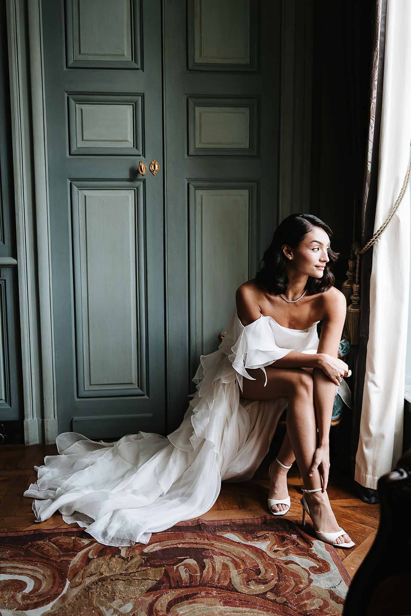 Bride in strapless ruffle gown seated by a tall window in a blue-paneled chateau room with herringbone floor