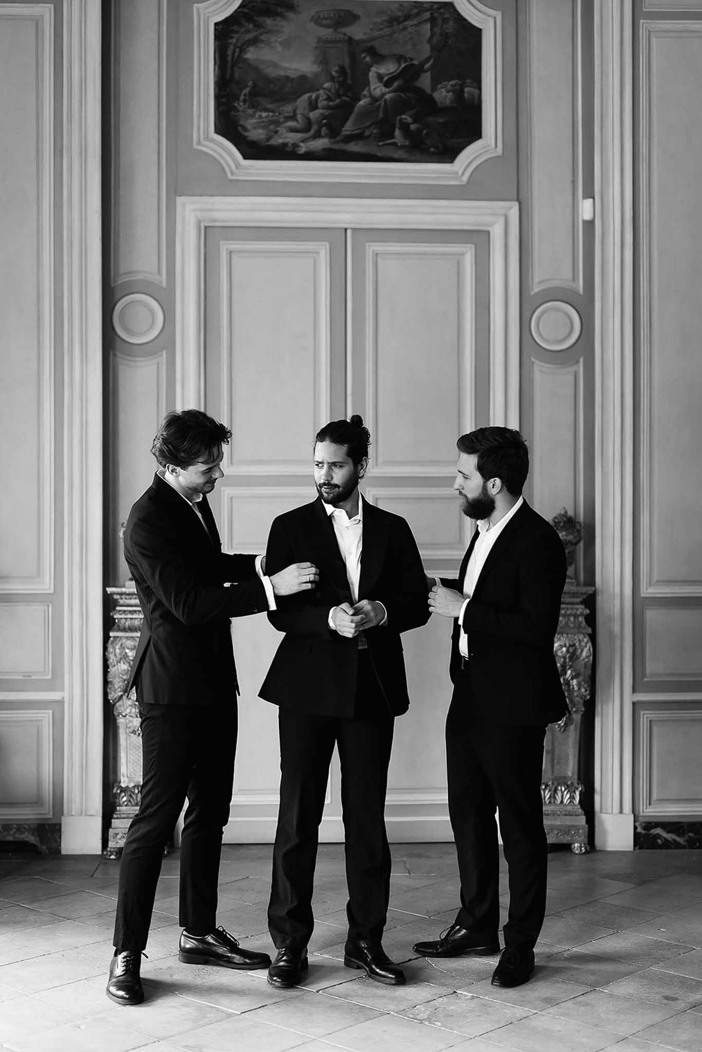 Black and white groom flanked by two groomsmen adjusting his suit before ornate boiserie paneling