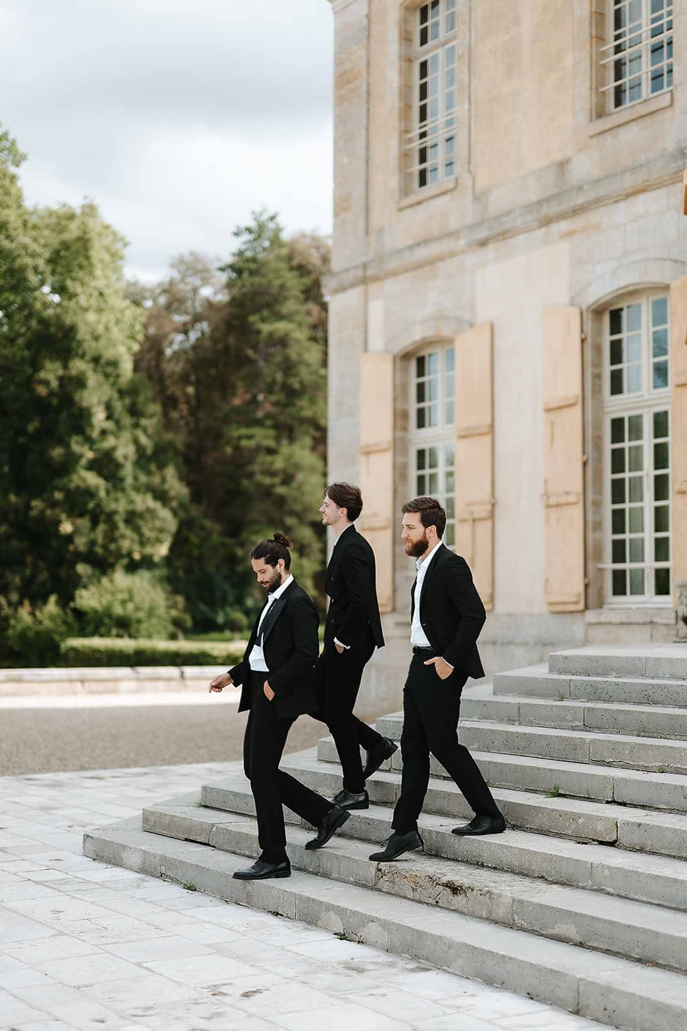 Three groomsmen in black suits walking down stone steps on exterior of French chateau