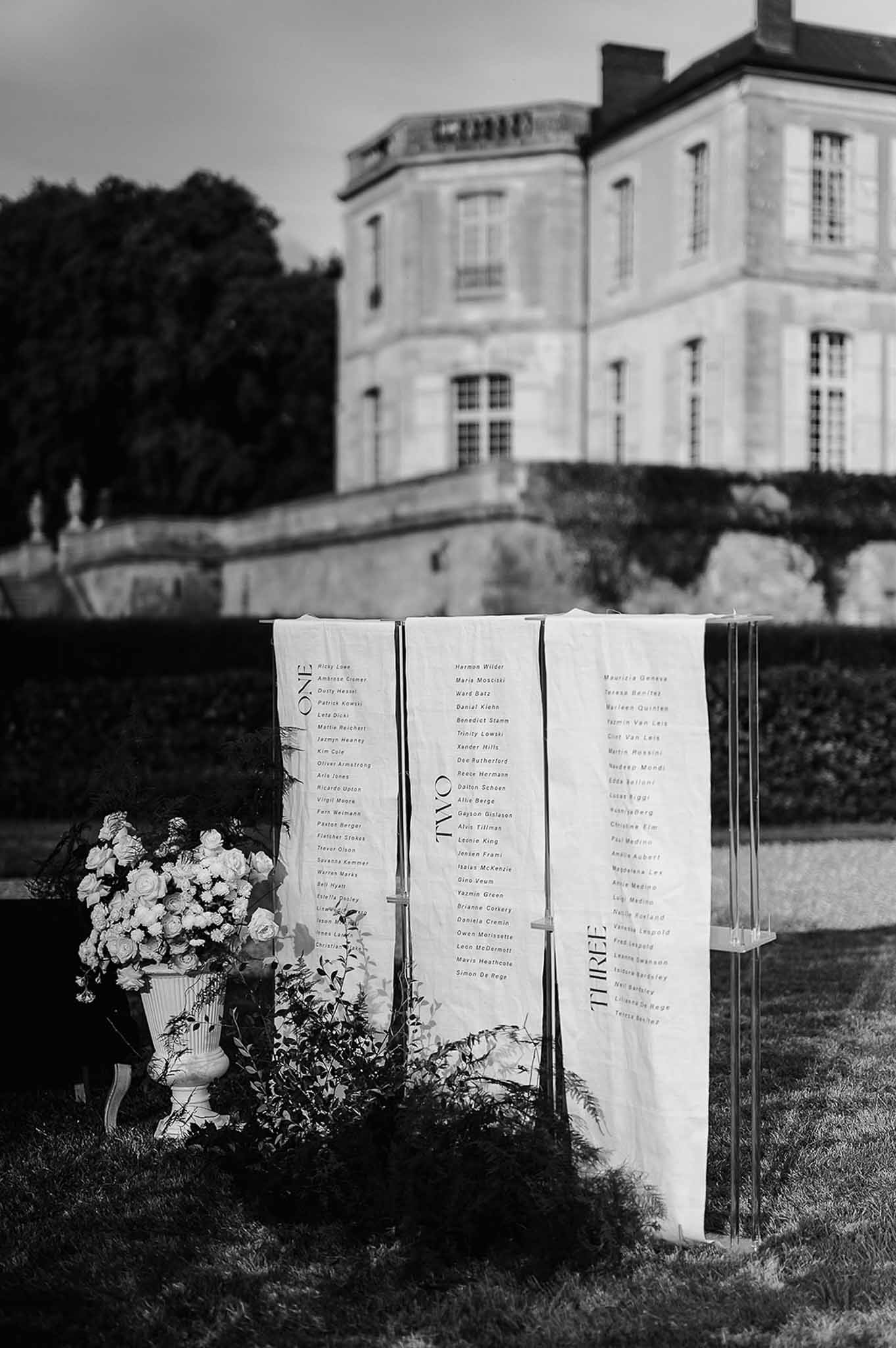 Black and white three-panel linen seating chart on acrylic frame with white rose urn before chateau on lawn