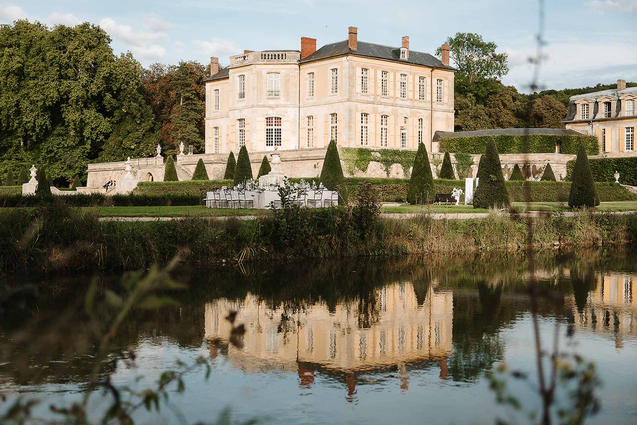 French chateau reflected in ornamental lake with formal topiary gardens and outdoor reception table on lawn