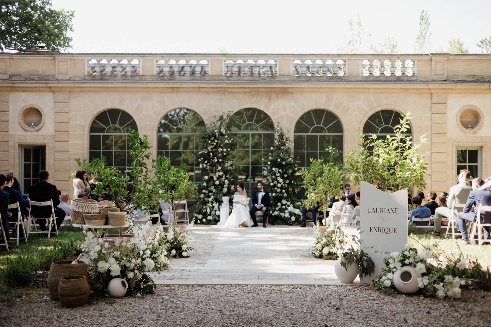 80 guests at orangery ceremony with circular white floral arch, rattan baskets, and sculptural welcome sign