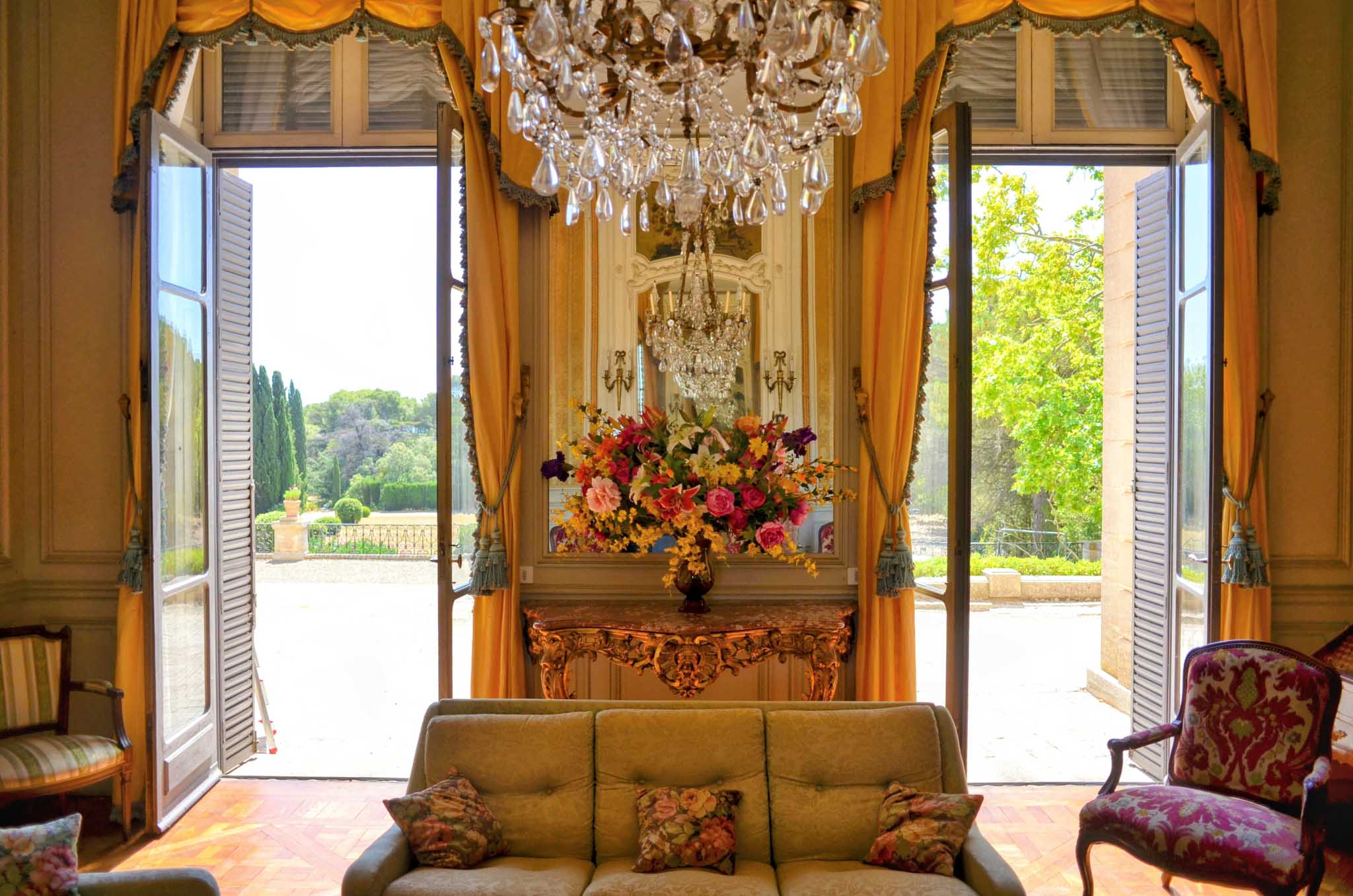 Grand chateau salon with crystal chandelier, gold drapes, Louis XV furniture, and French doors open to formal garden