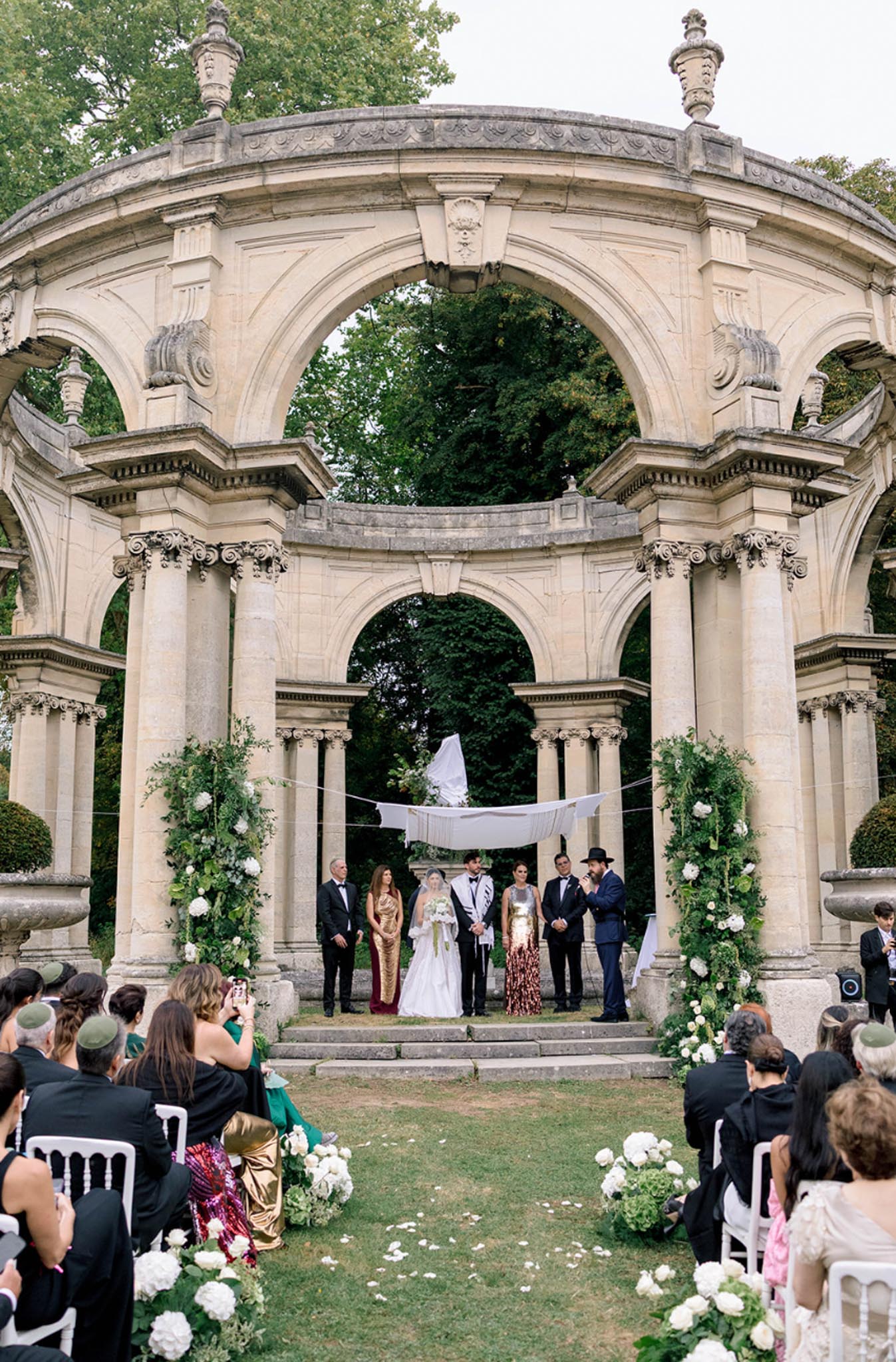 Jewish ceremony under white chuppah in stone rotunda with Corinthian columns white hydrangea arrangements and seated guests