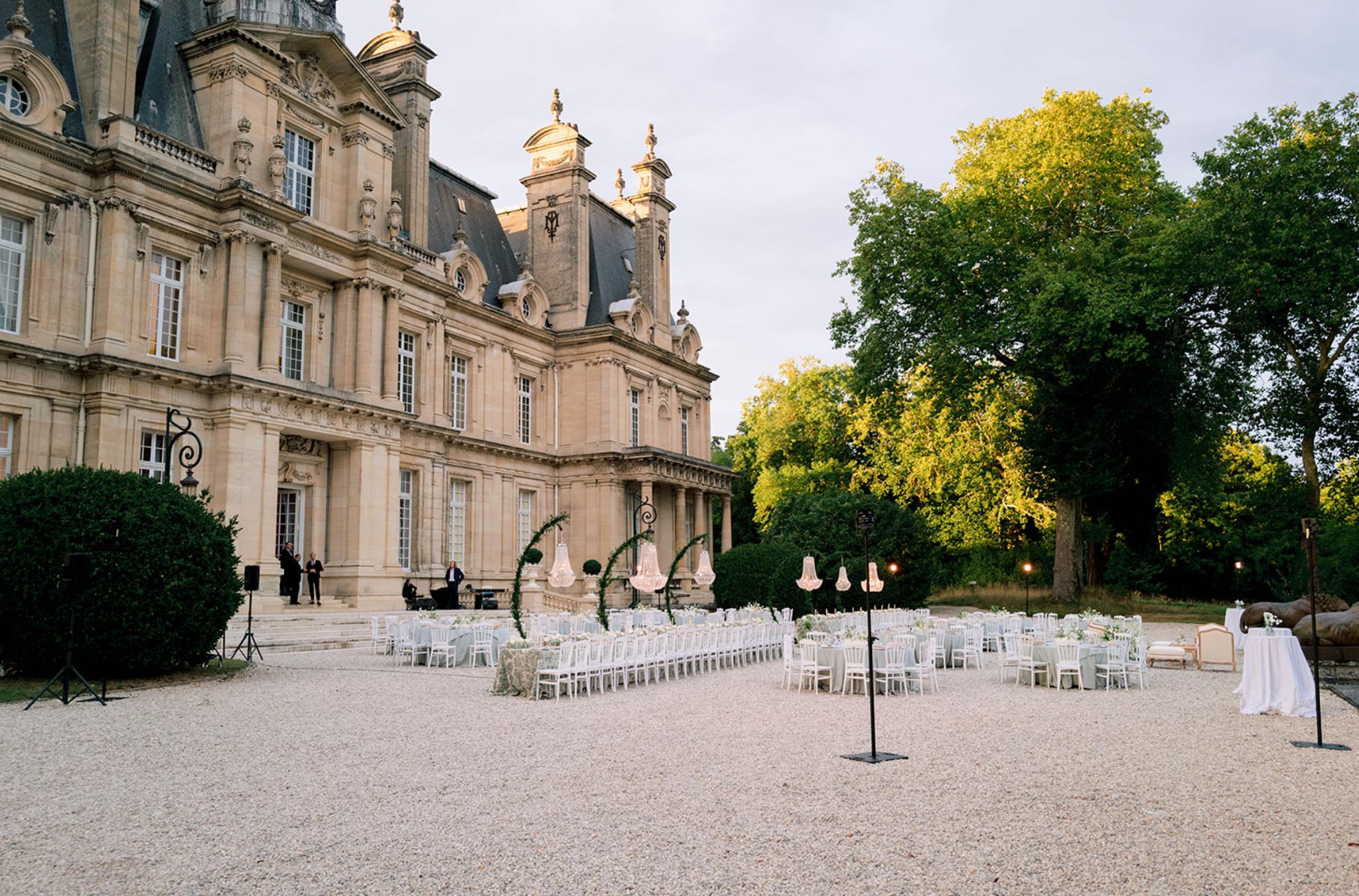 Dusk reception on chateau forecourt with long tables in blue-grey linens, crystal chandeliers, and greenery arches