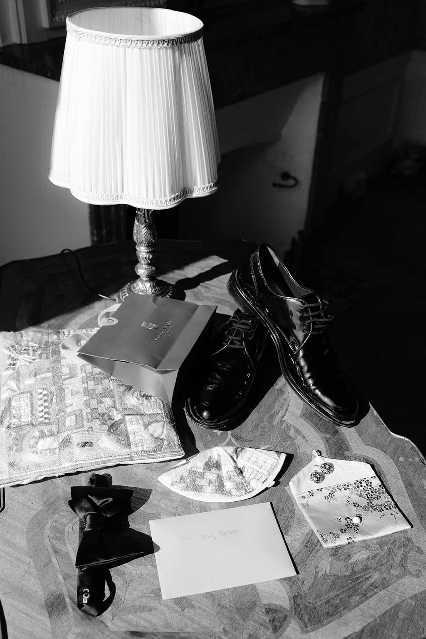 Black and white flat lay of groom accessories including Oxford shoes, bow tie, pocket square, and handwritten card