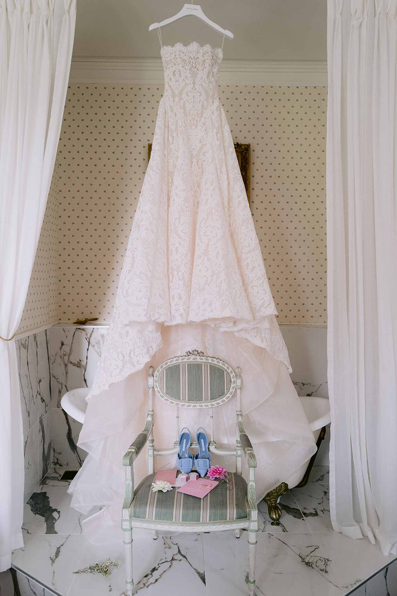 Blush pink lace wedding dress hung above sage green armchair with cornflower blue heels in French bathroom