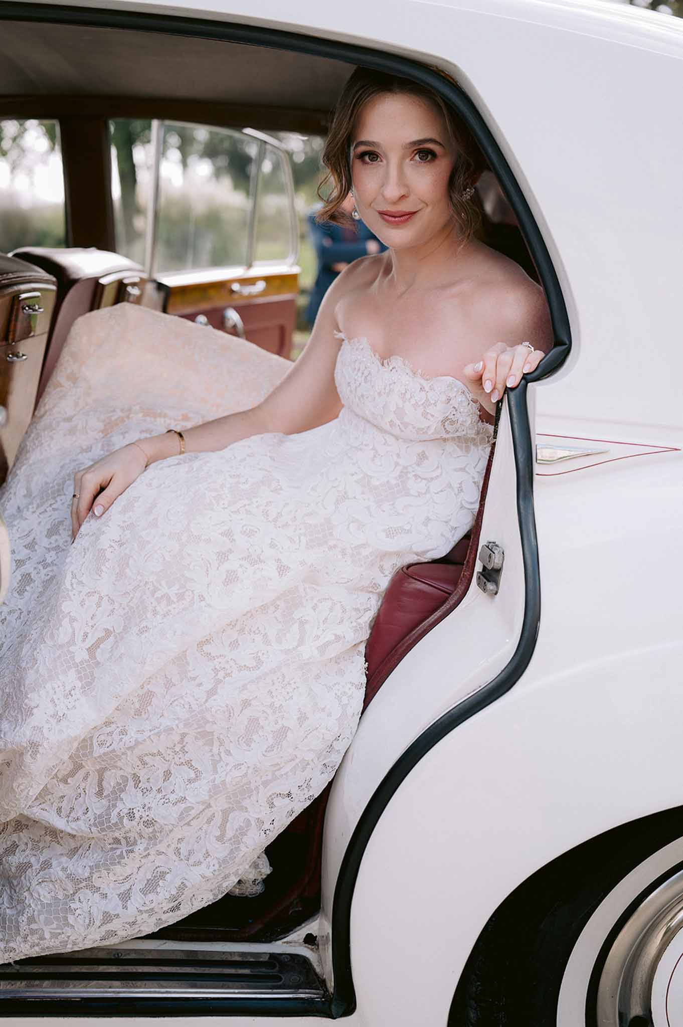 Bride in strapless floral lace gown with blush underlay seated in vintage white car with burgundy leather interior