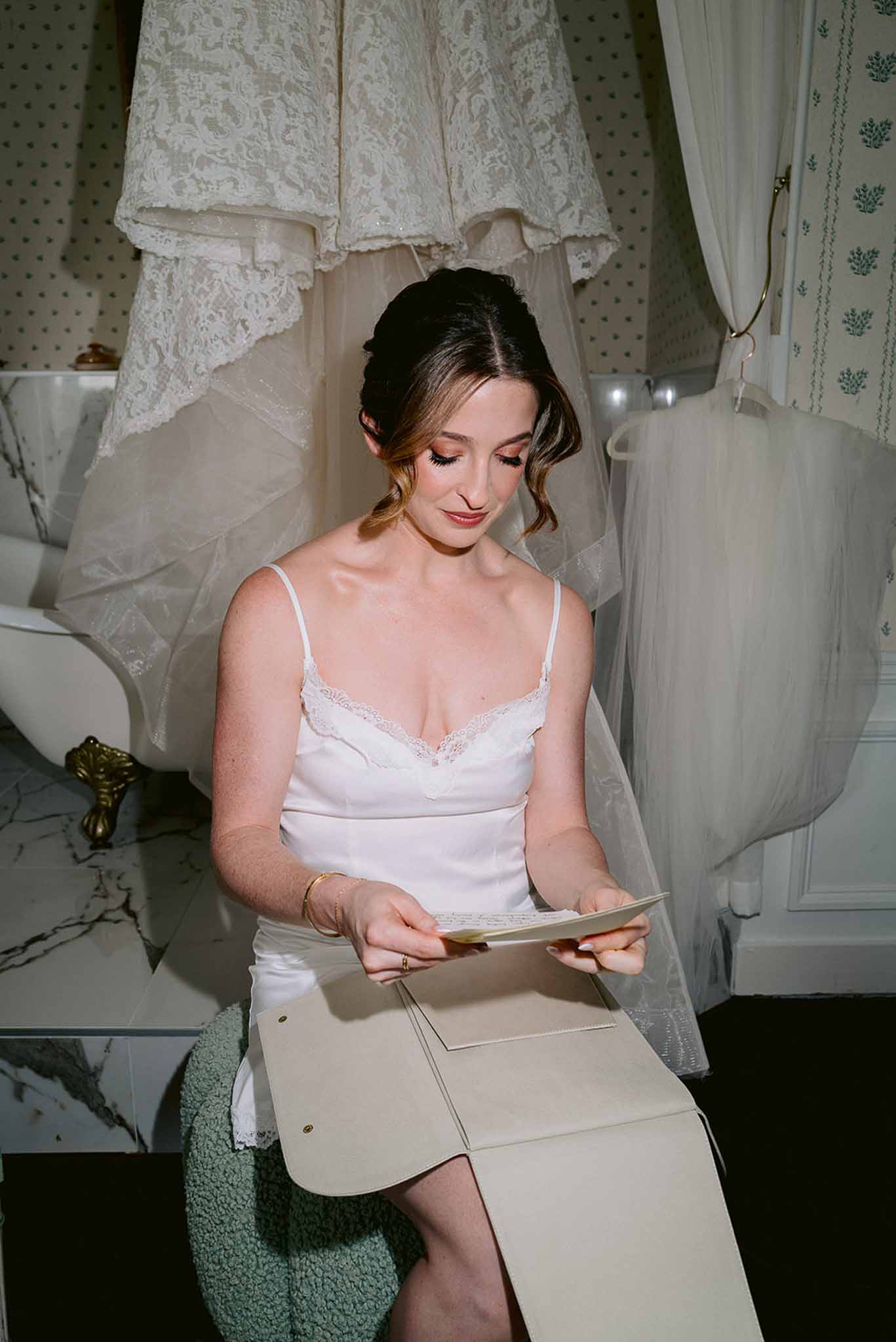 Bride reading handwritten letter during getting ready with wedding gown hanging behind her